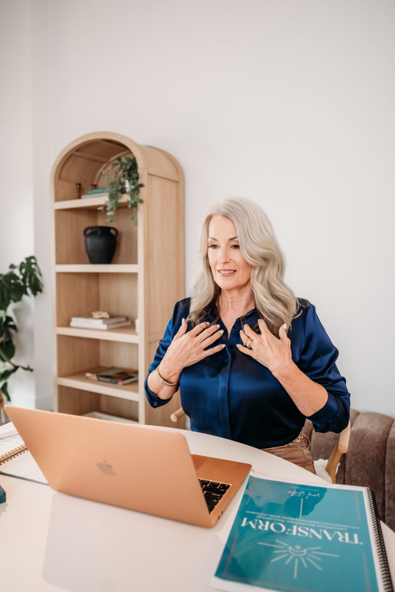 Woman with long gray hair smiling, sitting at a desk with a laptop and a book, in a bright room.