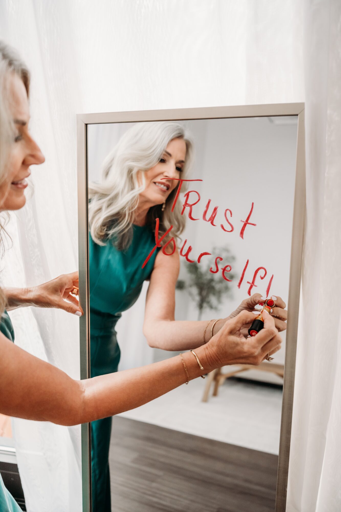 Woman writing on mirror with red marker, reflection visible, smiling, in bright room.