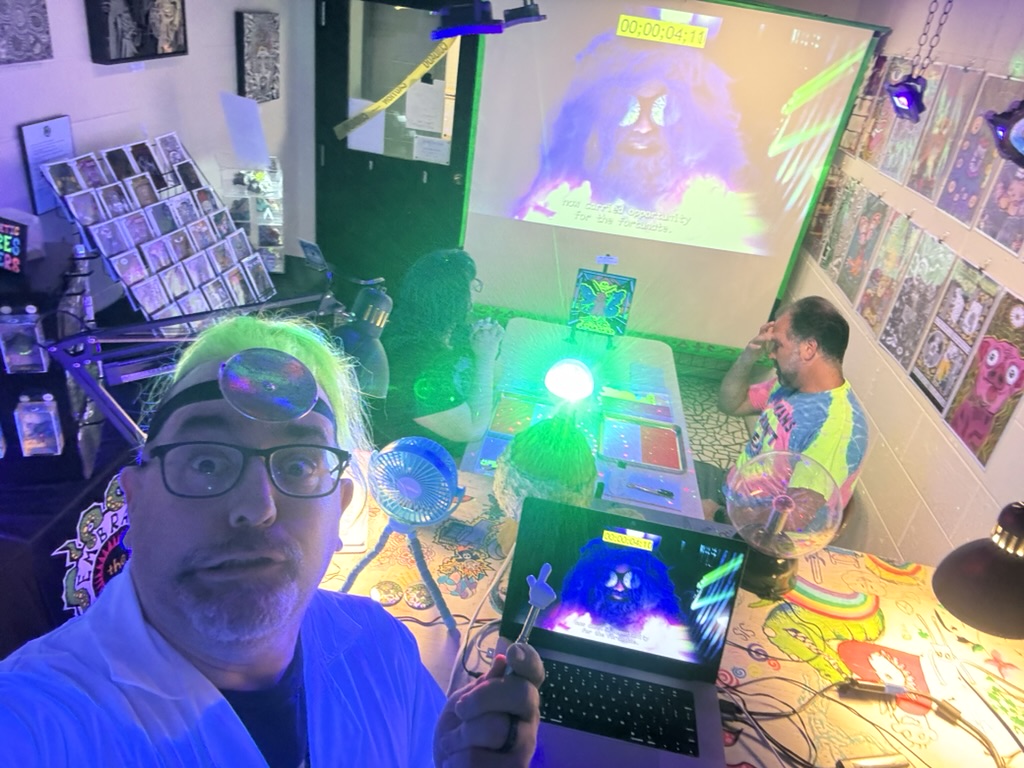 Man with glasses taking a selfie in a colorful room with a laptop, projector, and people playing games.