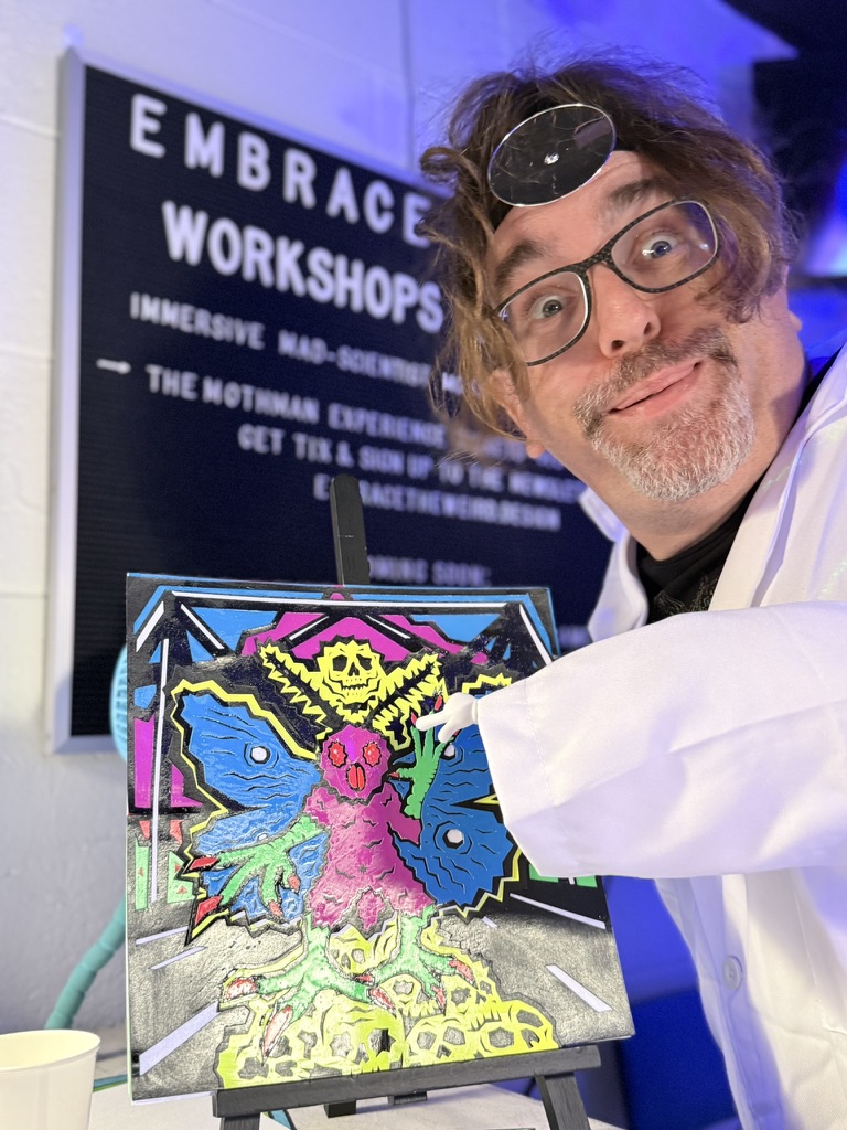 Person with glasses and curly hair in a white coat, smiling next to colorful artwork of a pink creature with blue butterflies, at workshop.