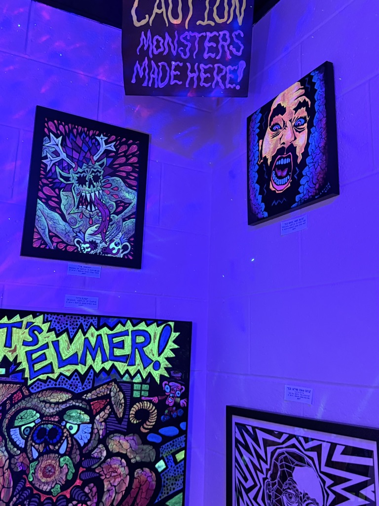 Three colorful artworks on a wall under purple lighting, including a skull, a face, and a cartoonish scene with text.