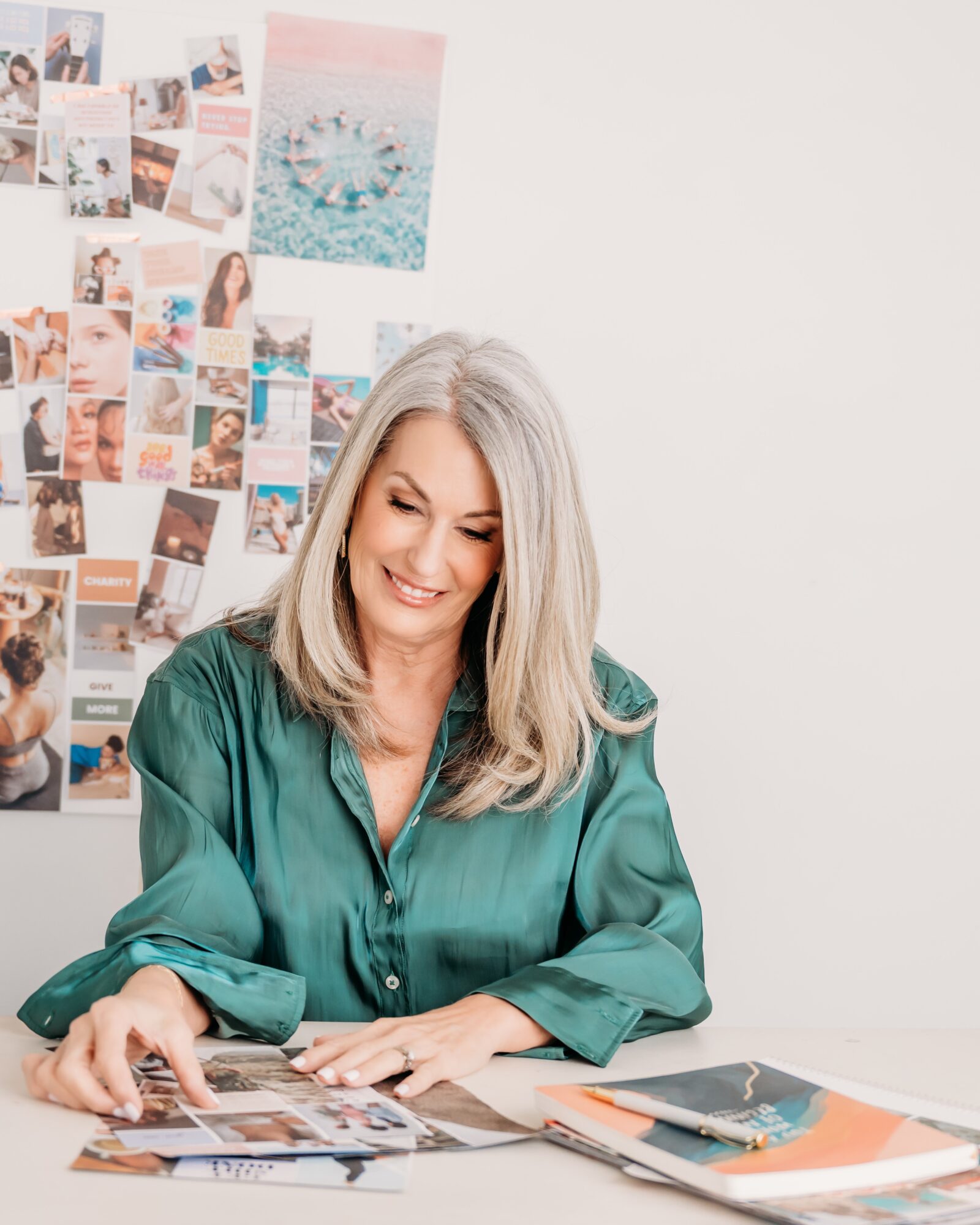 Woman with long gray hair smiling, sitting at a desk with magazines, in front of a wall with photos and artwork