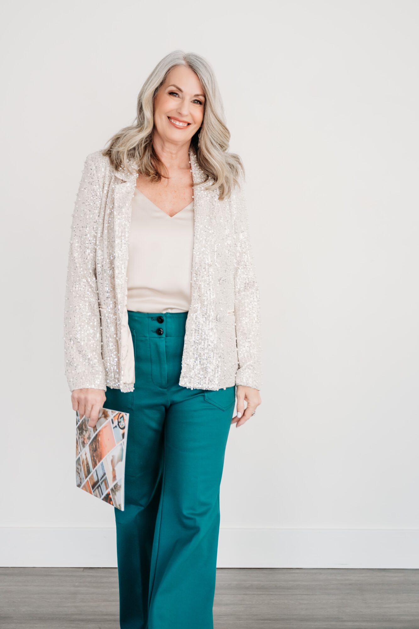 Smiling woman with gray hair in a white top, teal pants, and a light cardigan, holding a magazine, standing against a plain background.