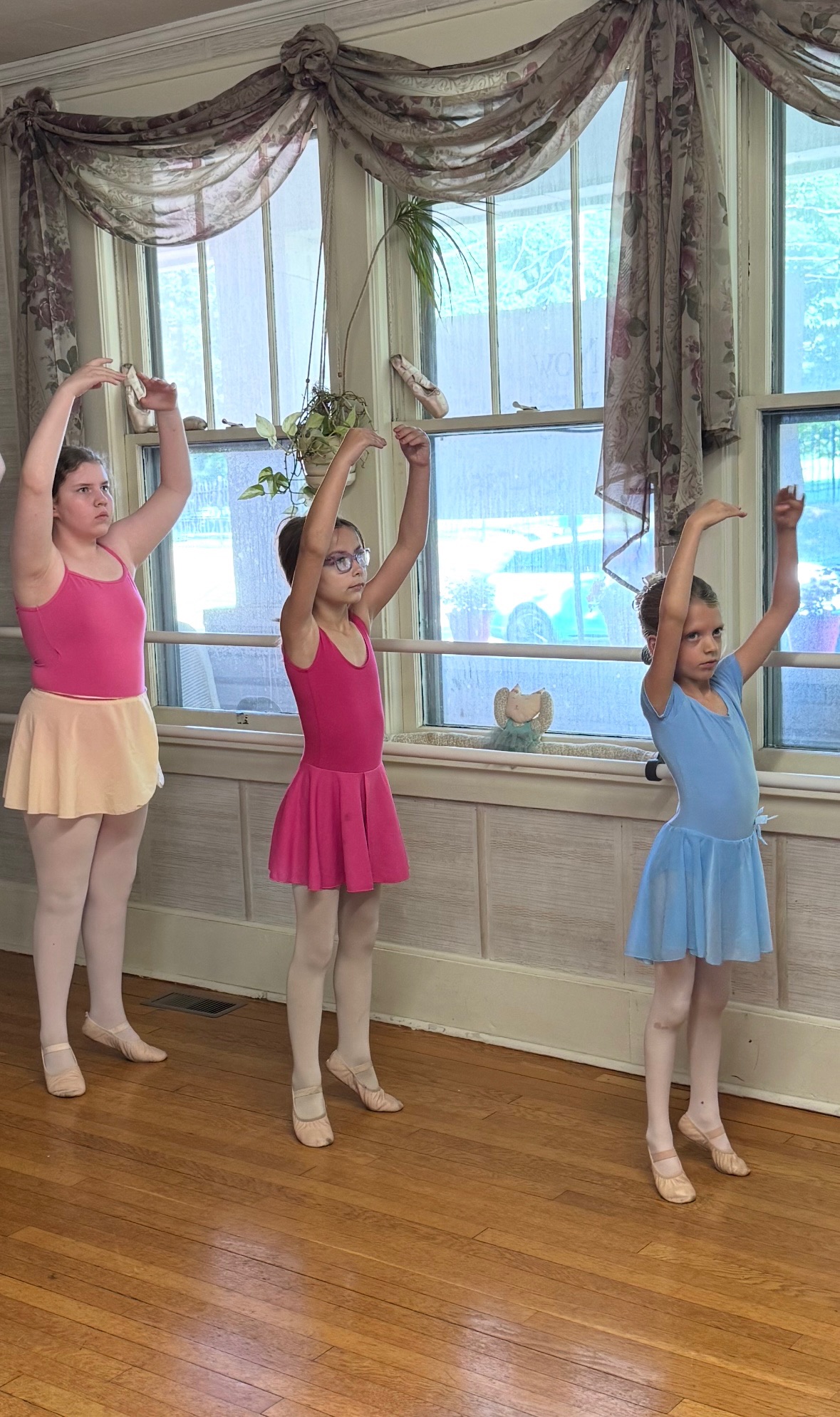 Three people in dresses raise their arms while standing in front of tall windows.