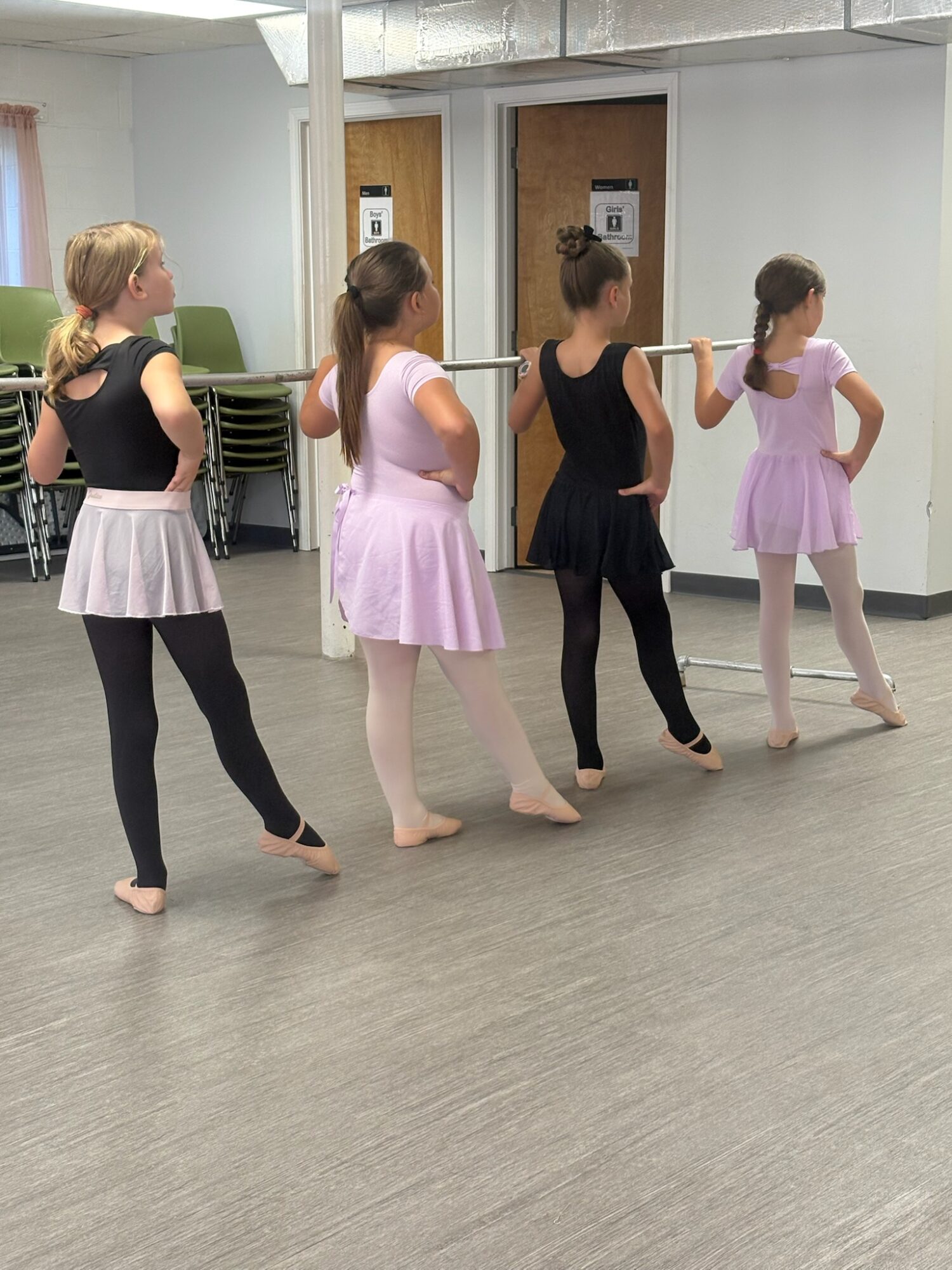 Four people in dancewear stand at a ballet barre in a studio.