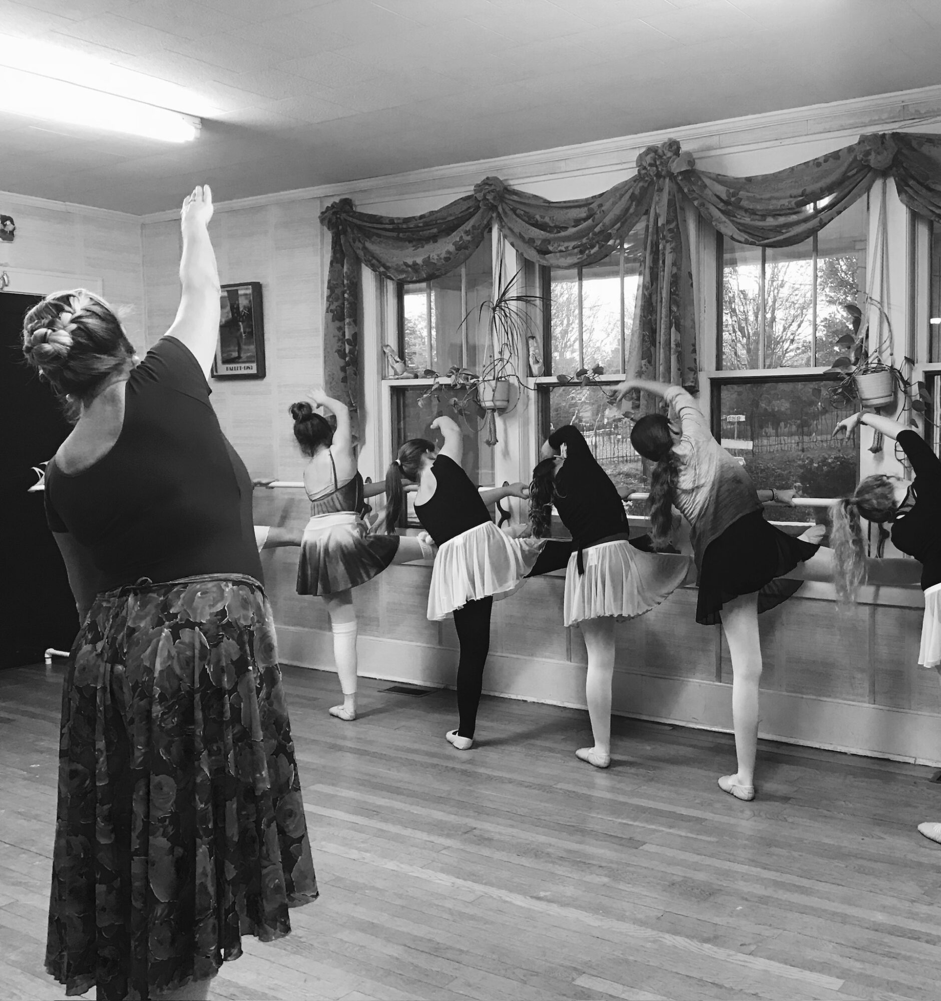 A group of dancers stretch at a barre while an instructor raises one arm in a studio.