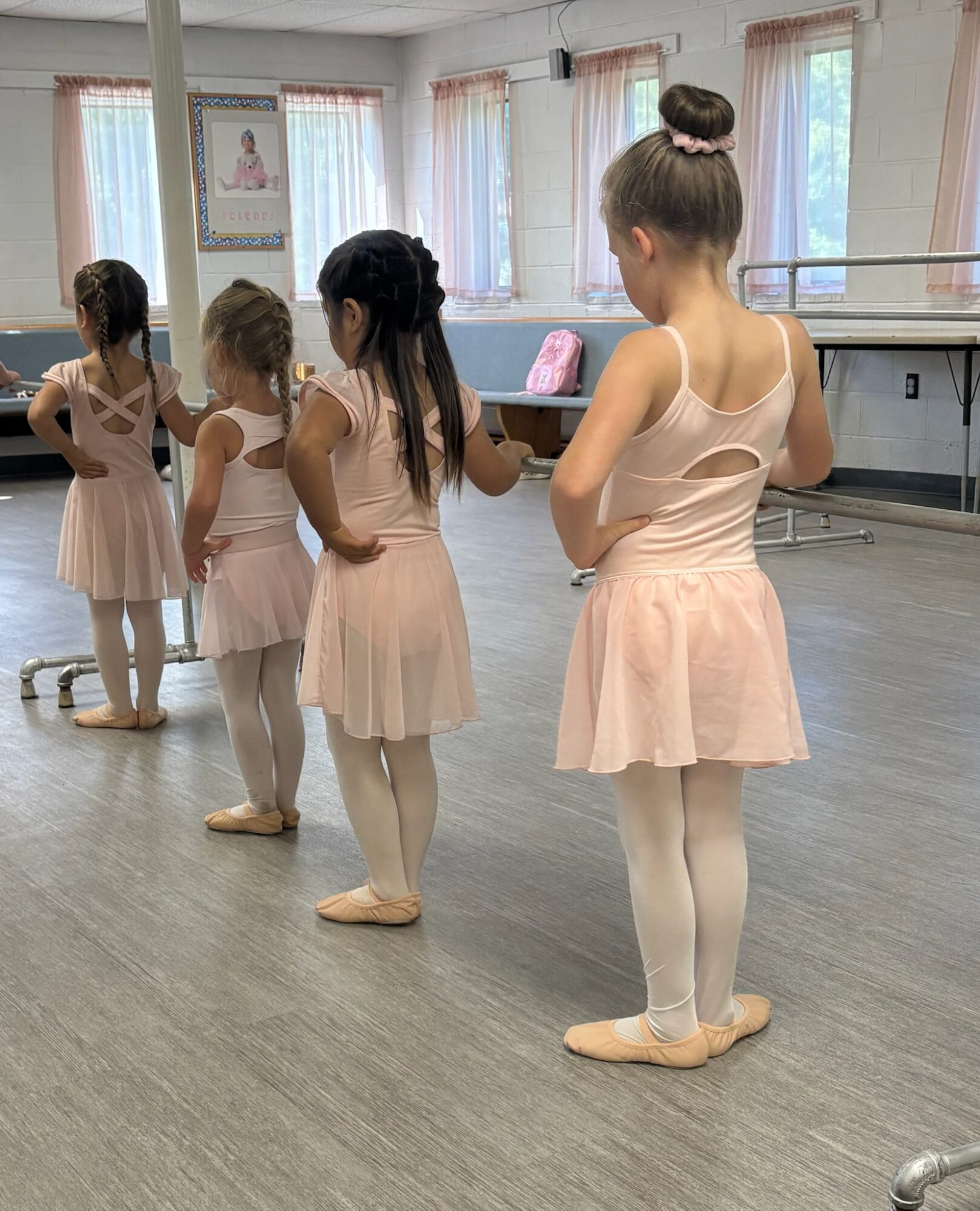 Four ballet students in pale pink leotards and skirts stand in a dance studio.