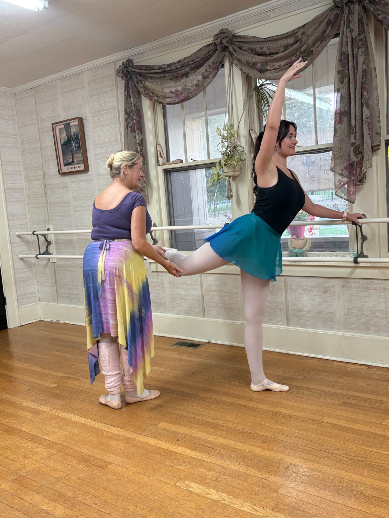 A person balances on one leg with the other extended behind, assisted by another person in a dance studio.