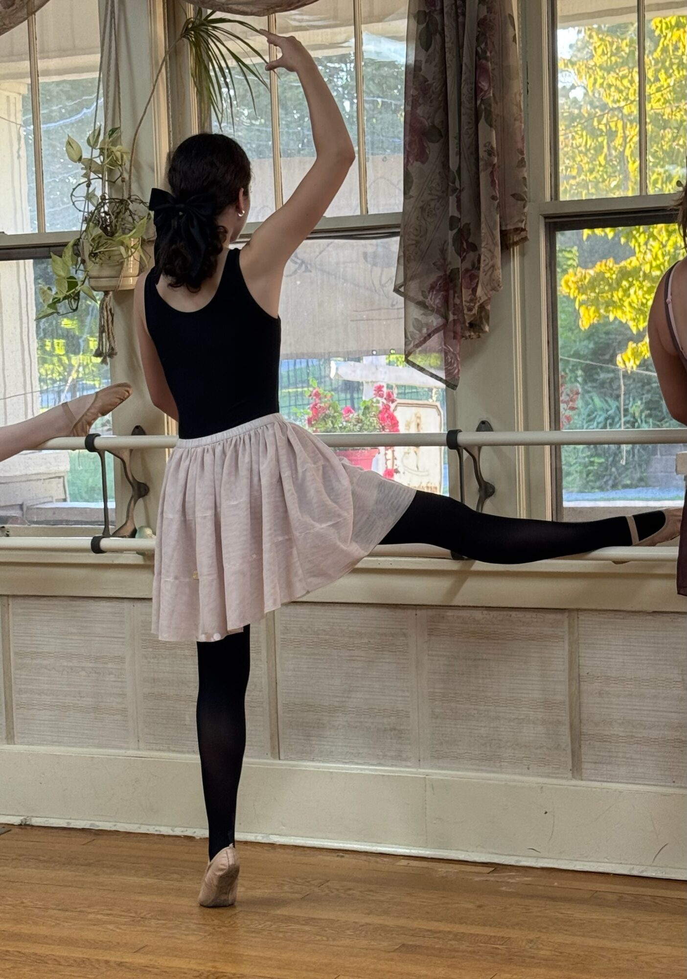 A person in a black top and pale skirt stretches one leg along a ballet barre in a room with large windows.