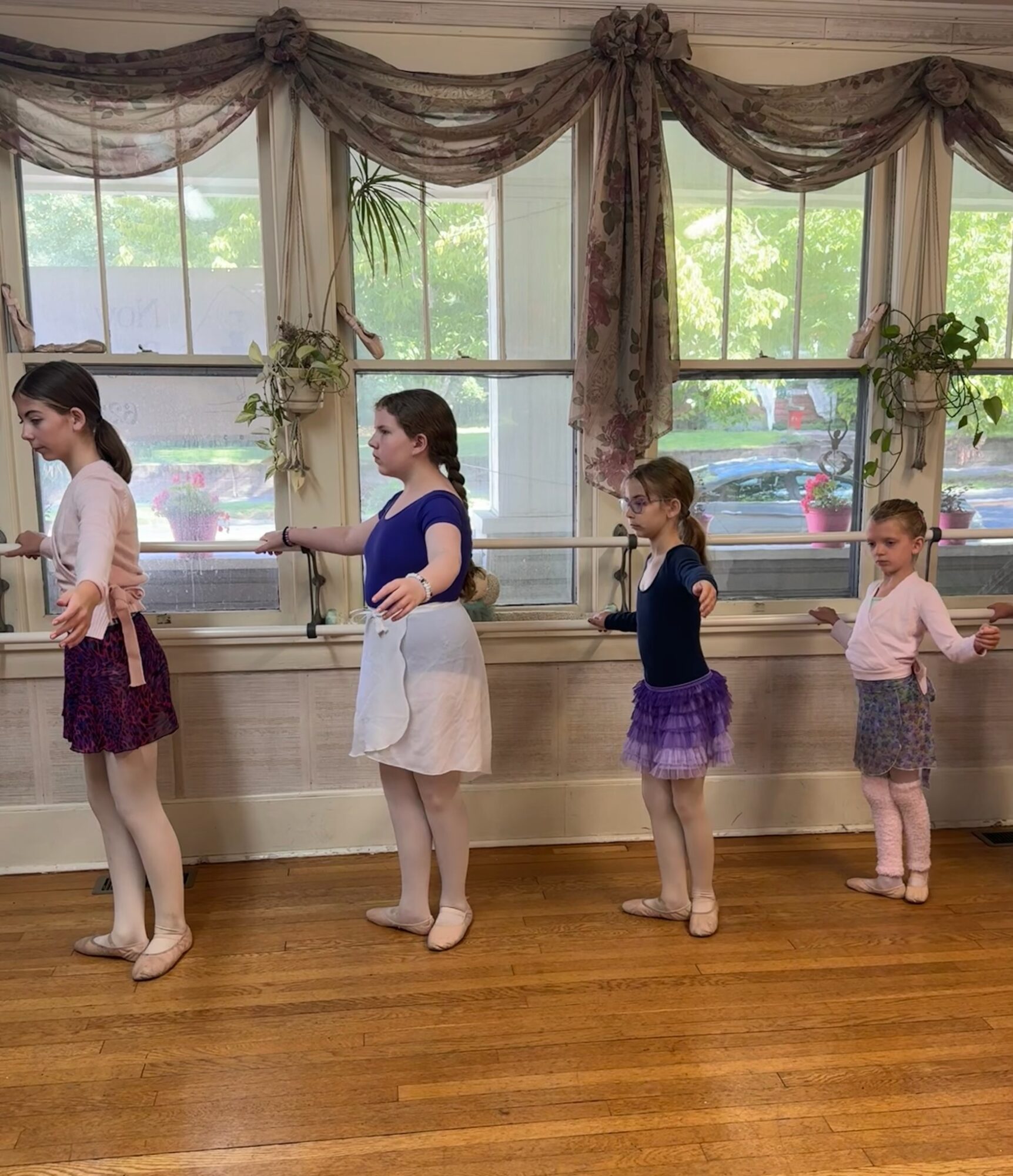 Four people in ballet attire stand in a line at a barre in a dance studio.
