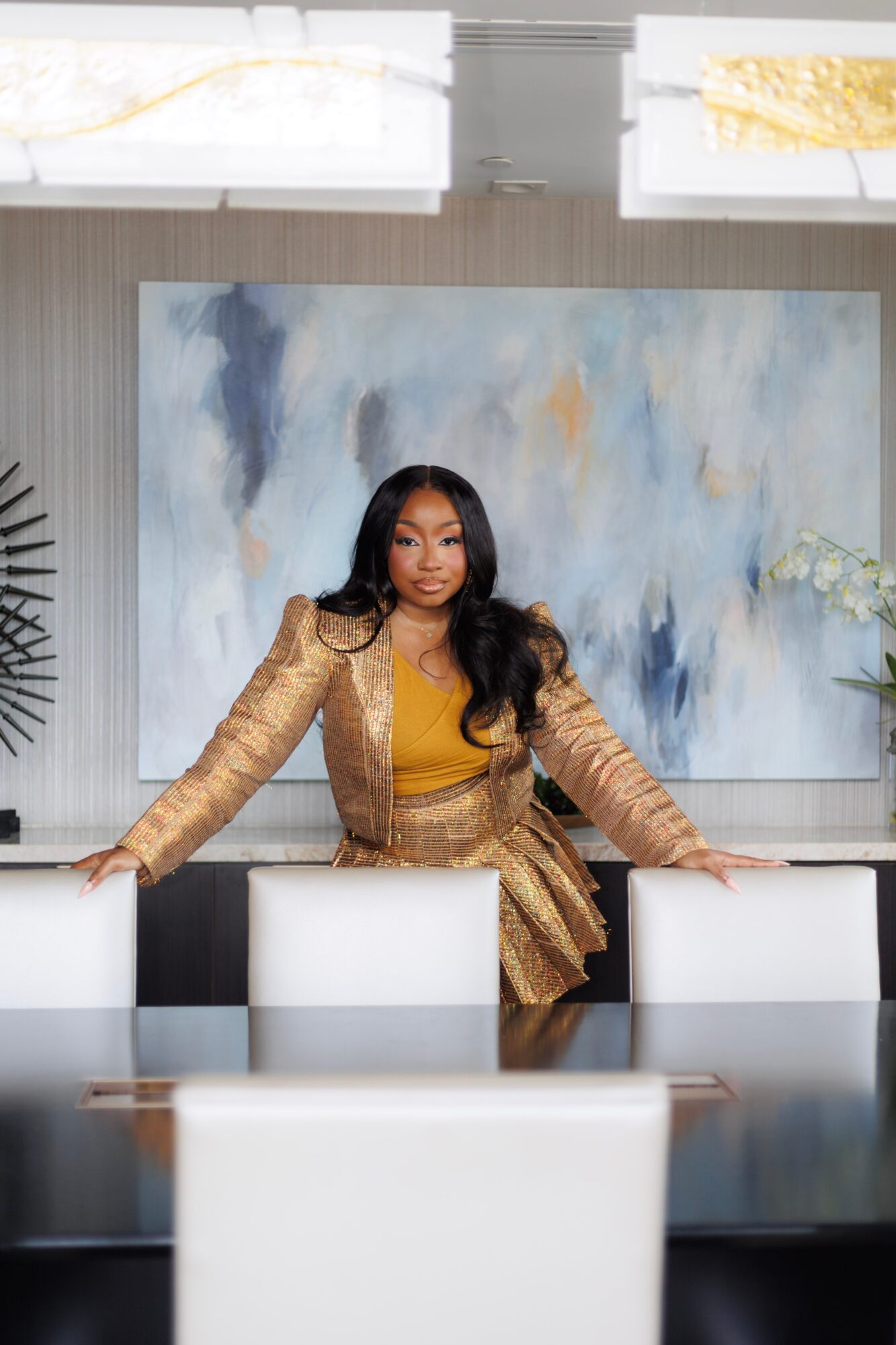 Woman in gold jacket and dress standing behind a conference table in a modern room with abstract painting.