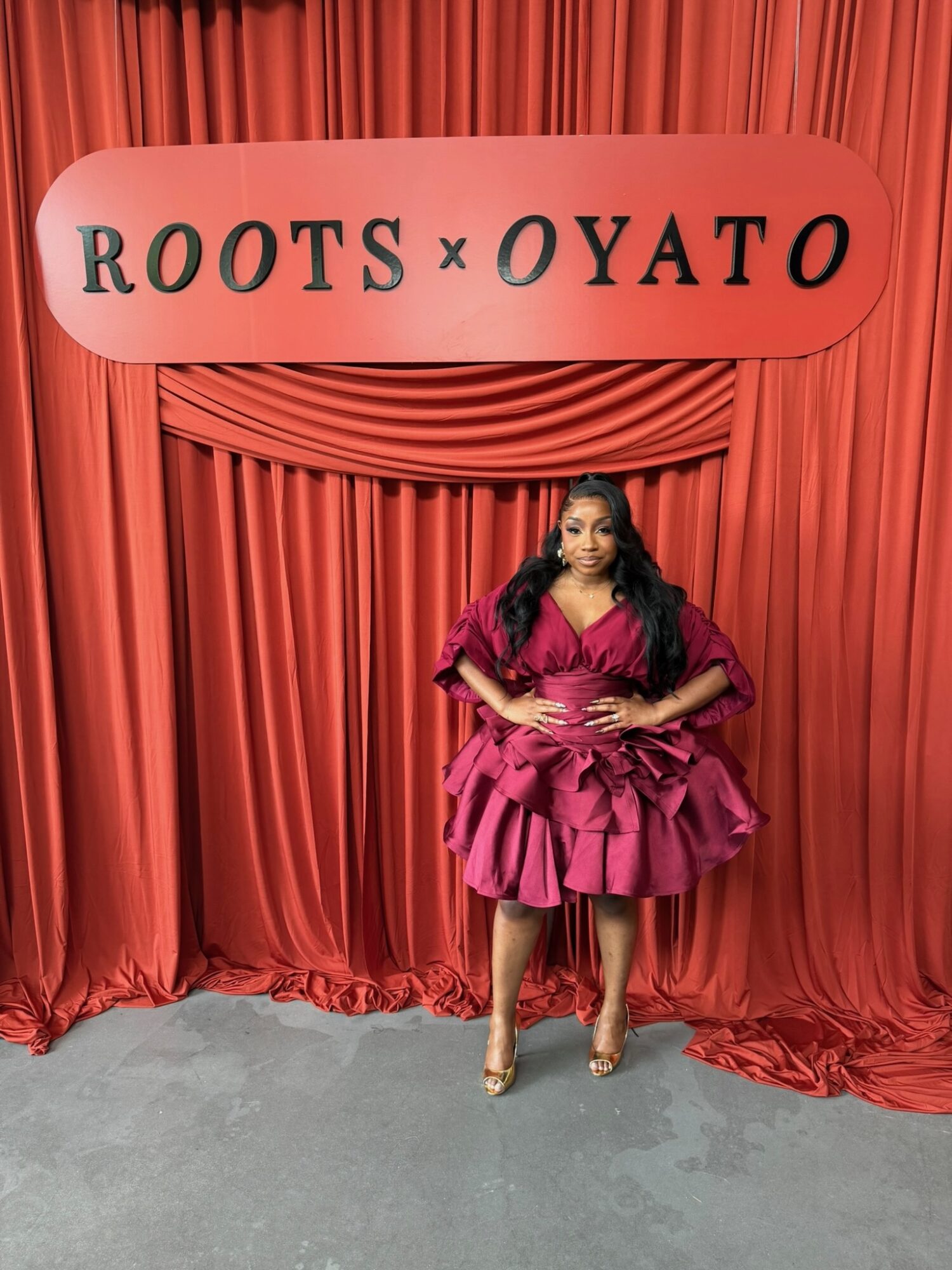 Woman in pink dress standing in front of red curtain with a sign reading 'ROOTS x OYATO'.