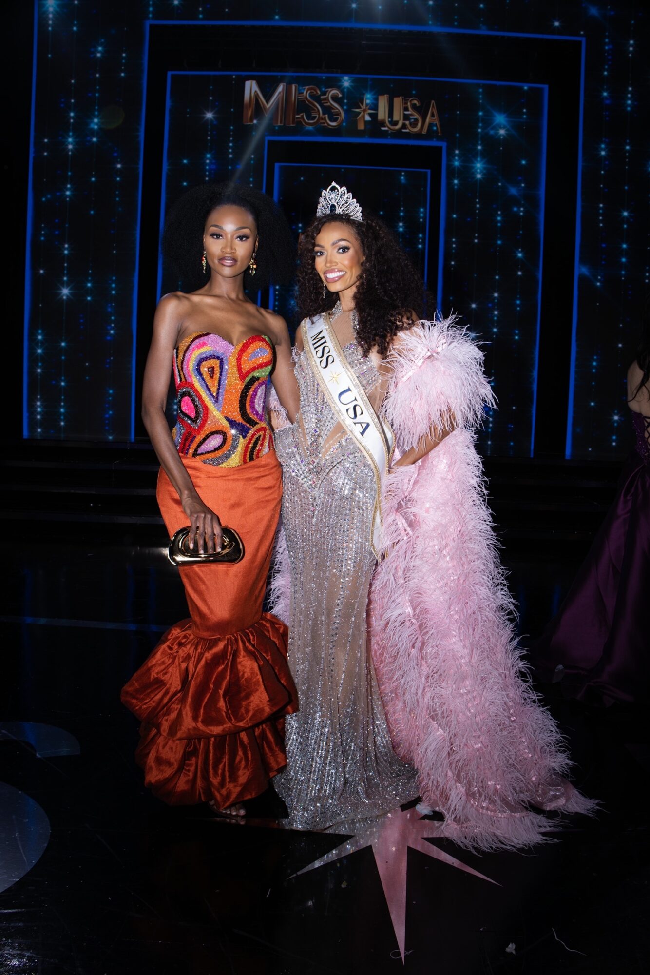 Two women in evening gowns on stage, one wearing a crown and sash, with a star on the floor, background displays 'Miss USA'.