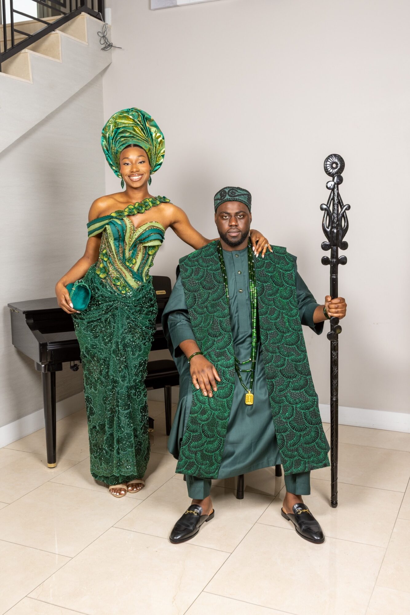 A woman in colorful traditional attire and a man in green clothing pose indoors, with a piano and decorative staff in the background.