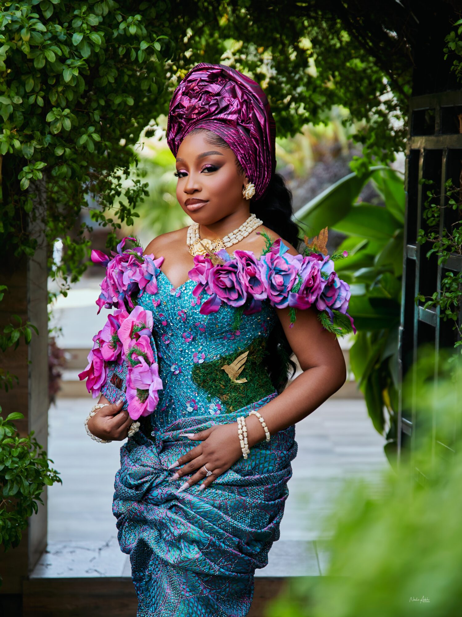Woman in traditional attire with purple headwrap and floral embellishments, standing outdoors among greenery.
