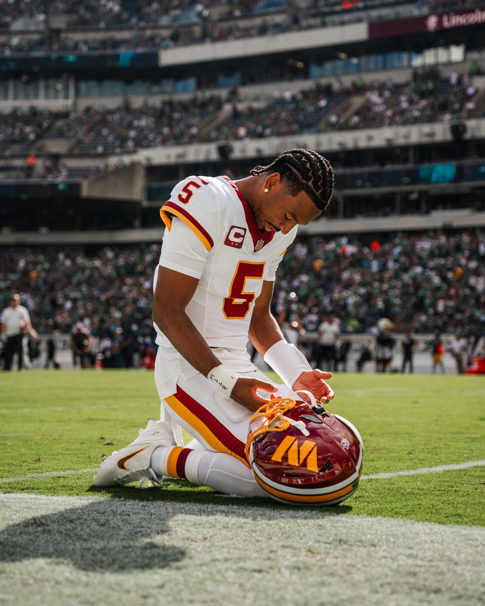 Football player kneeling on field with helmet, in uniform, head bowed, stadium with spectators in background