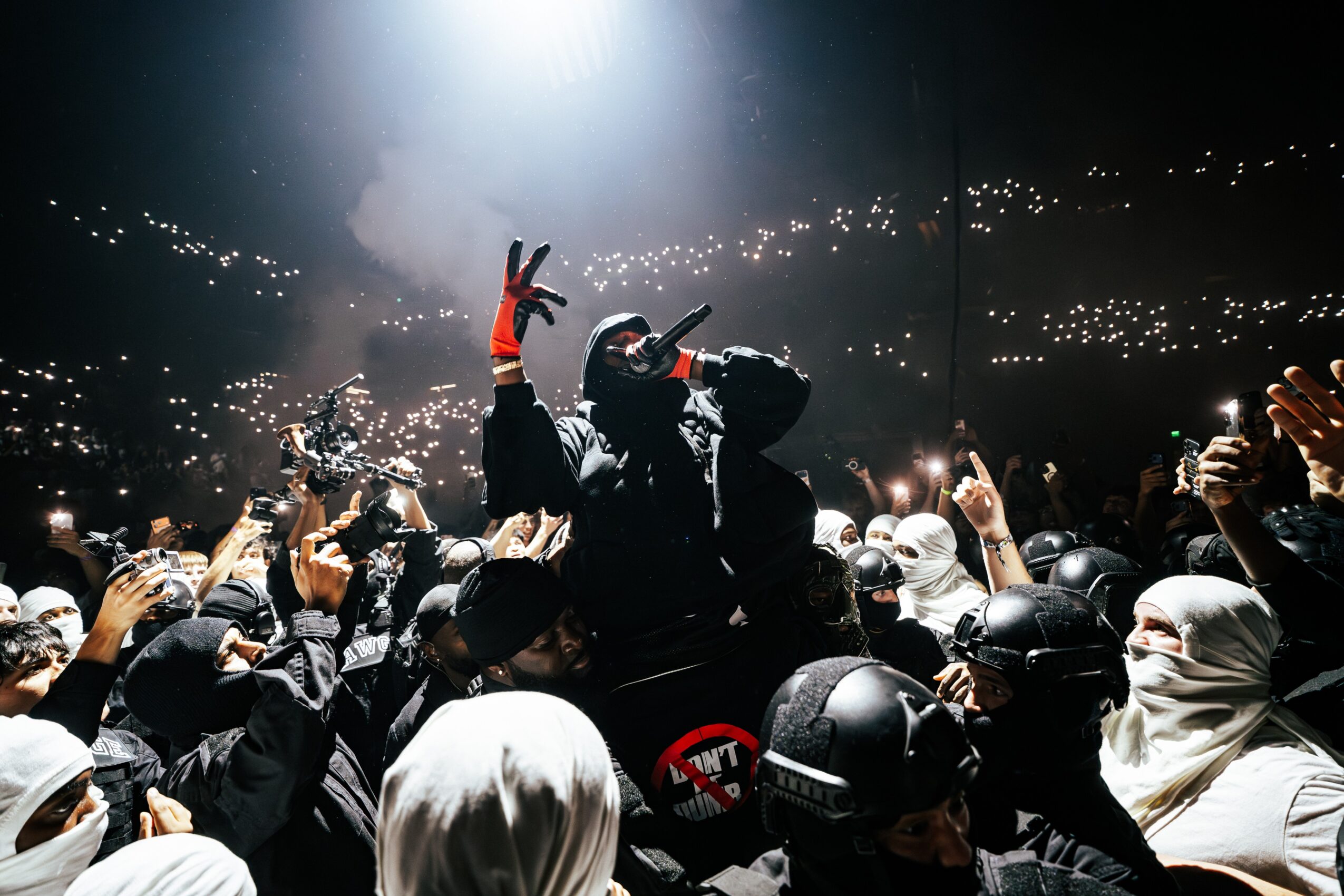 Crowd at concert with a performer on stage, some people holding up peace signs, in a dark venue with lights.