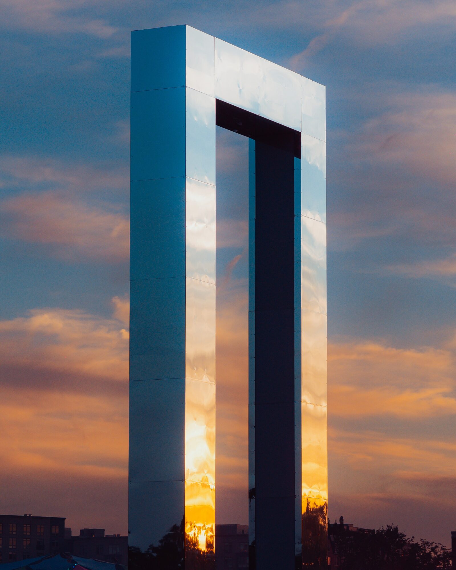 Modern metallic arch structure reflects the sunset sky, with a city skyline and trees in the background.