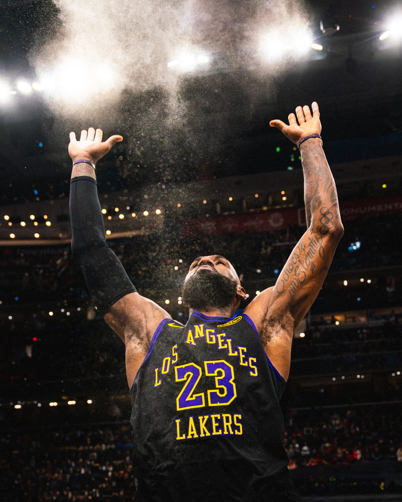 Basketball player wearing Los Angeles Lakers jersey with number 23, raising arms in celebration, dust or powder in the air, stadium lights overhead.