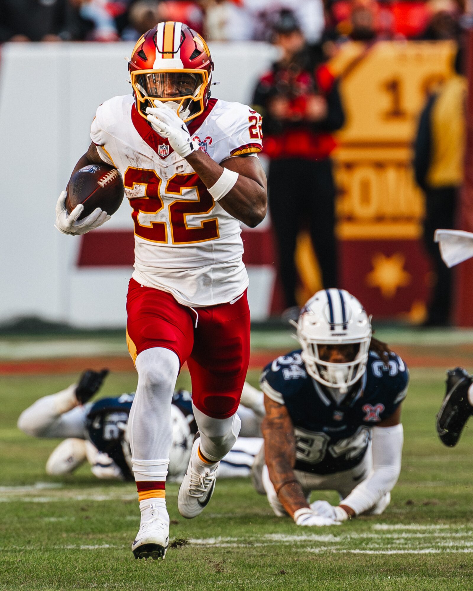 Football player in white and red uniform running with ball, opposing player on ground, blurred background