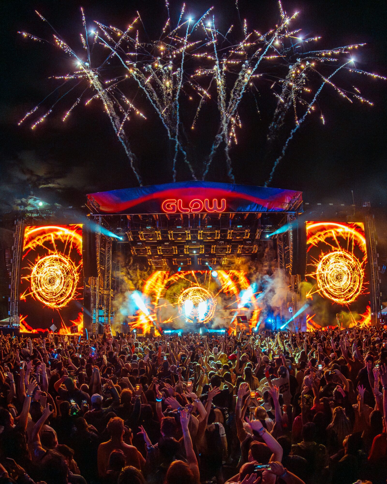Large crowd at outdoor concert with fireworks in the night sky, stage with bright lights and visuals, and a sign reading 'GLOW'.
