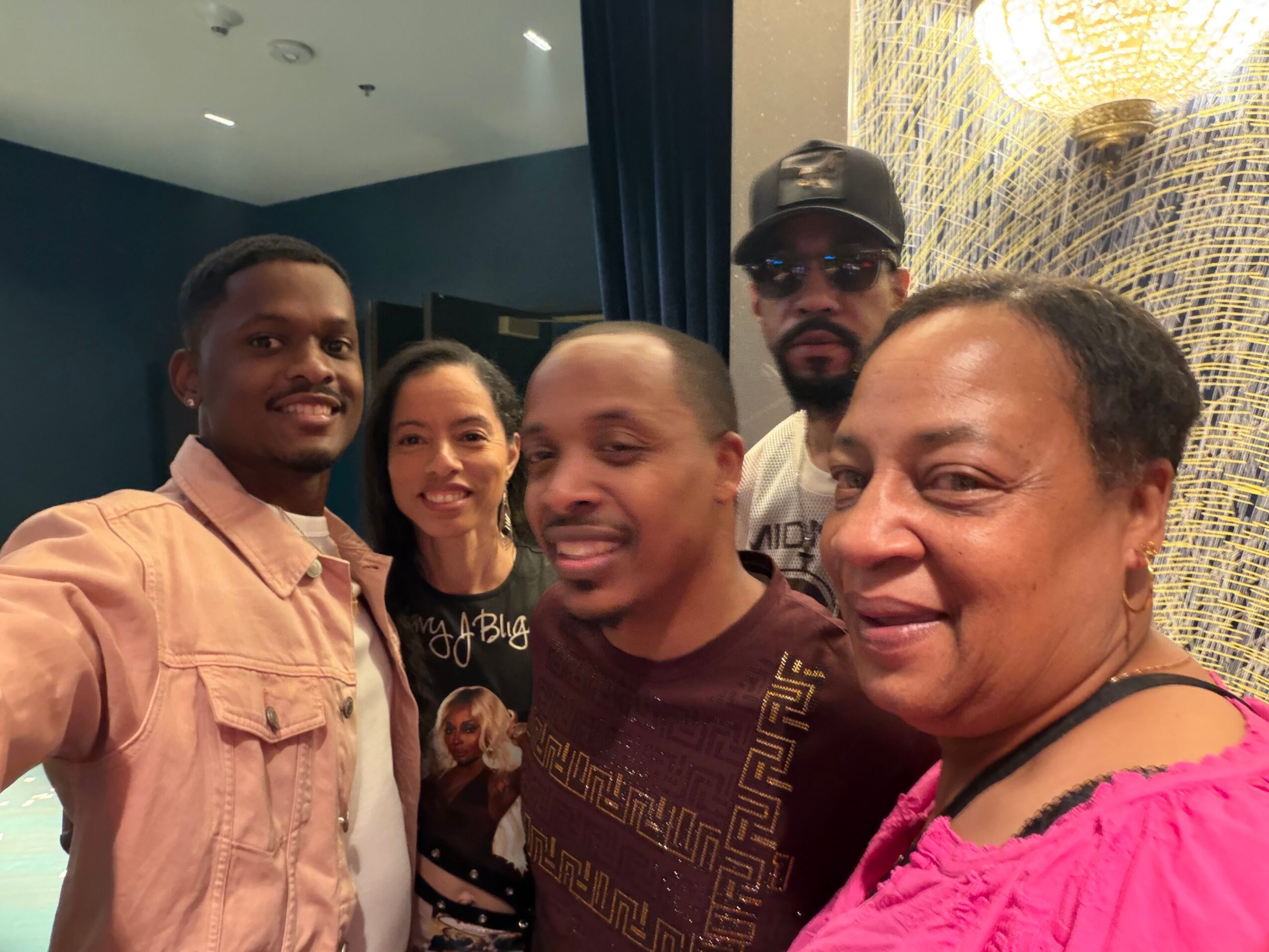 Five people smiling and posing for a photo indoors, some with their heads close together, background with a curtain and ceiling lights.