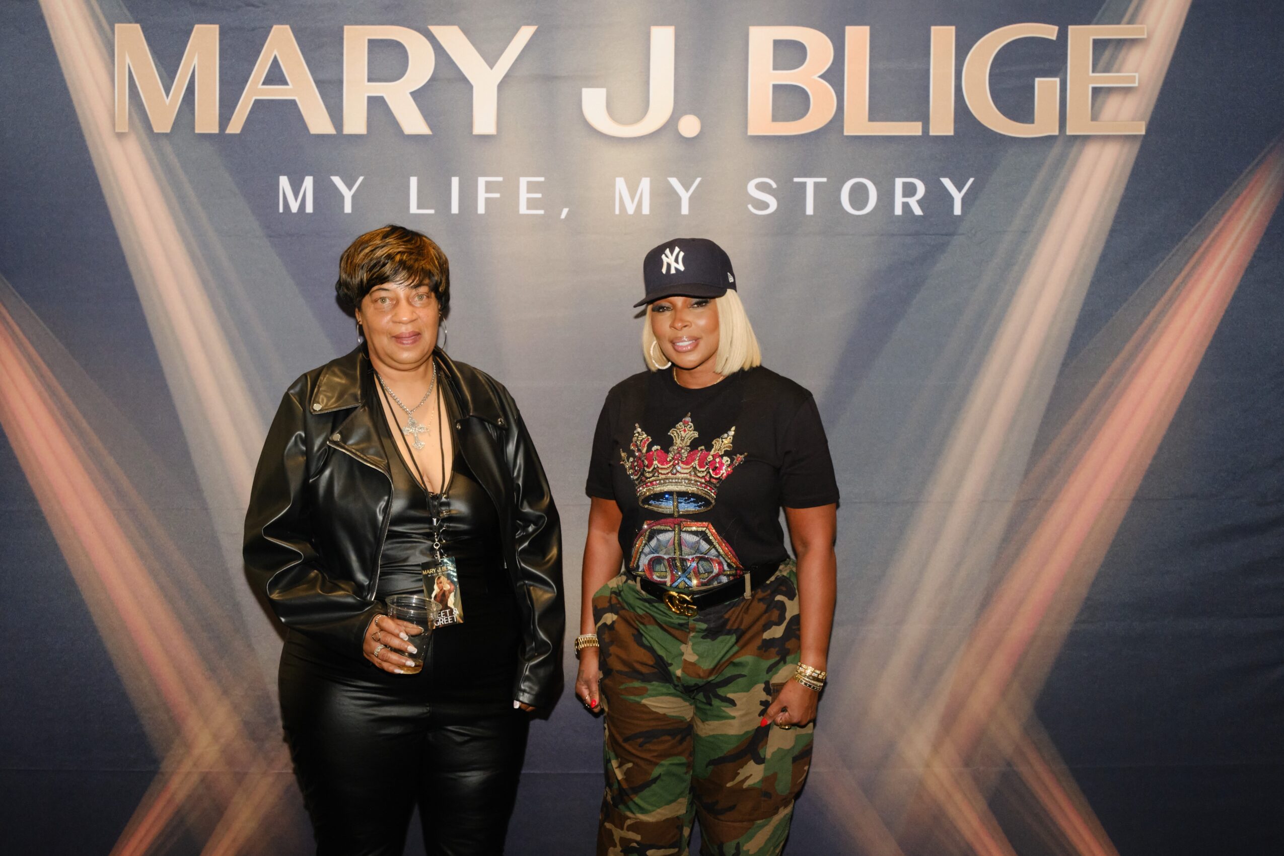 Two women standing in front of a backdrop with text, one holding a drink, both smiling.