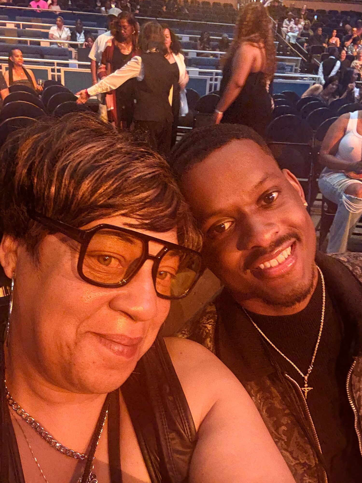Two smiling women taking a selfie at an outdoor event, with people and seating in the background.