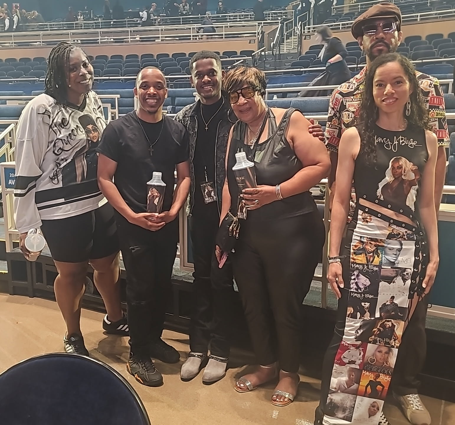 Group of six people standing together in an indoor venue, smiling, holding drinks, with empty seats in the background.