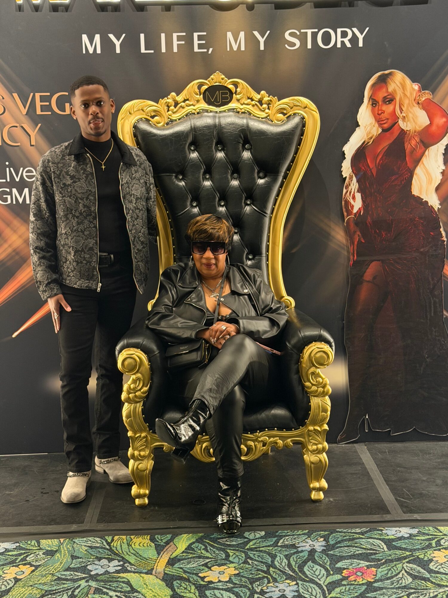 Person sitting on a large ornate black and gold throne with two people standing beside and a poster in the background.