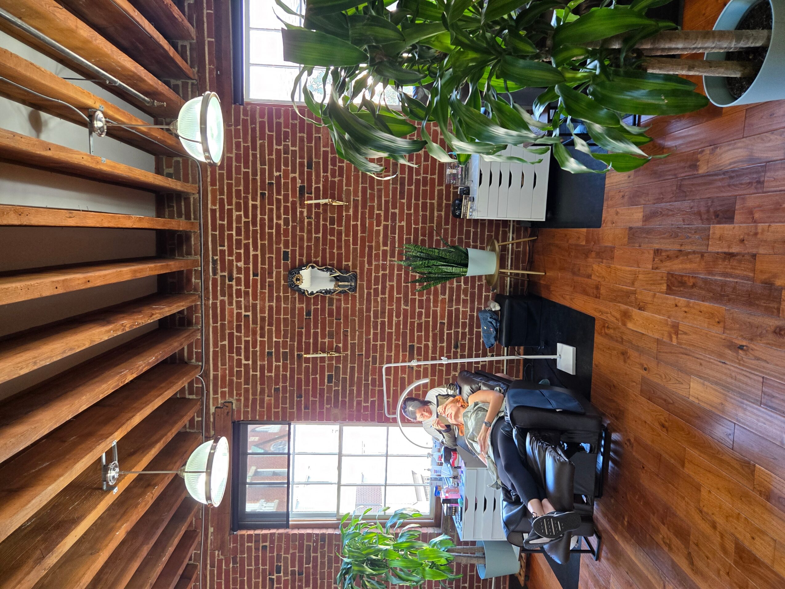 Person sitting in a black chair in a room with brick wall, wooden ceiling, large window, and plants.