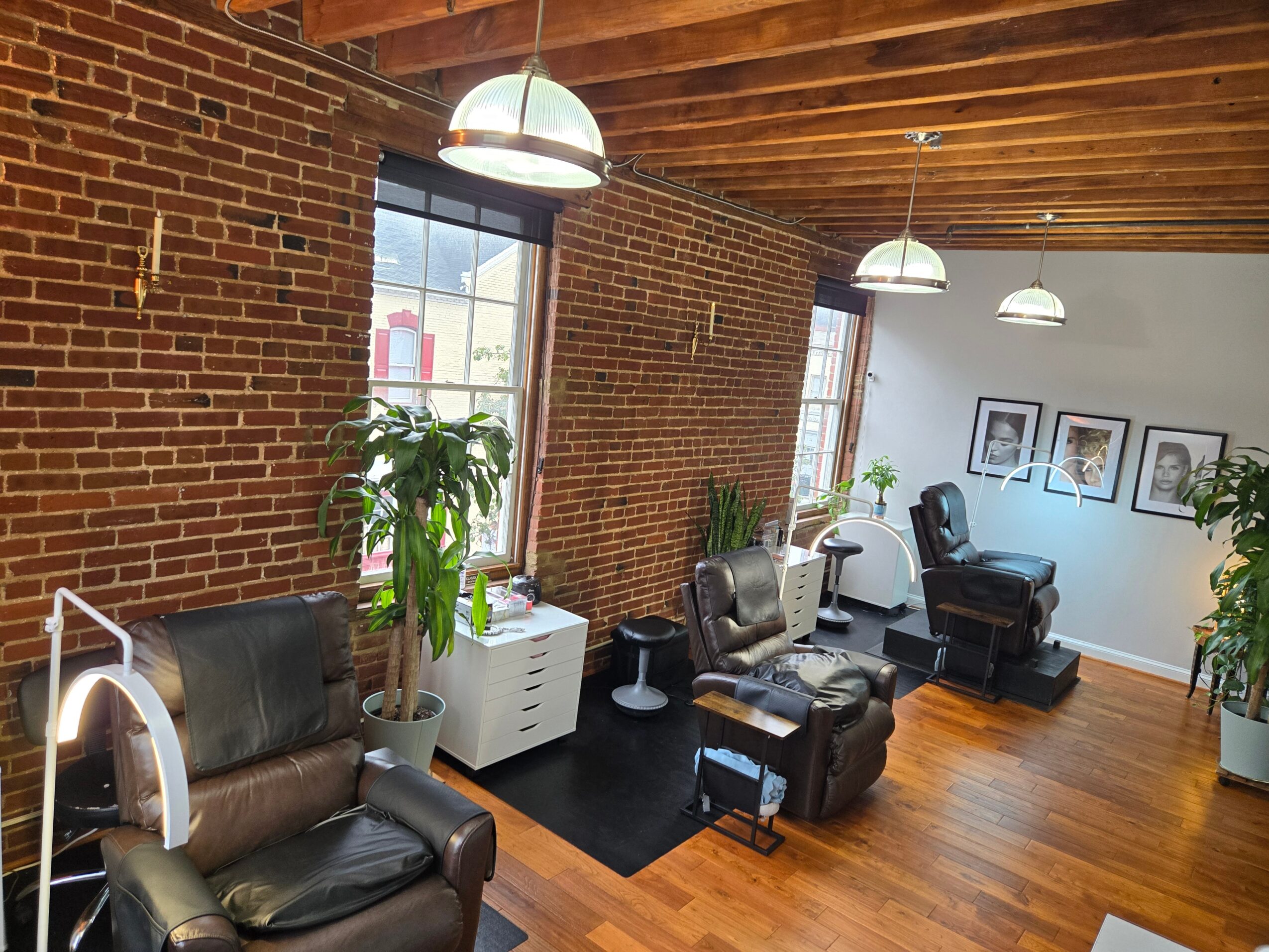 Interior of a room with brick walls, wooden ceiling, large windows, leather chairs, plants, and framed pictures on the wall.
