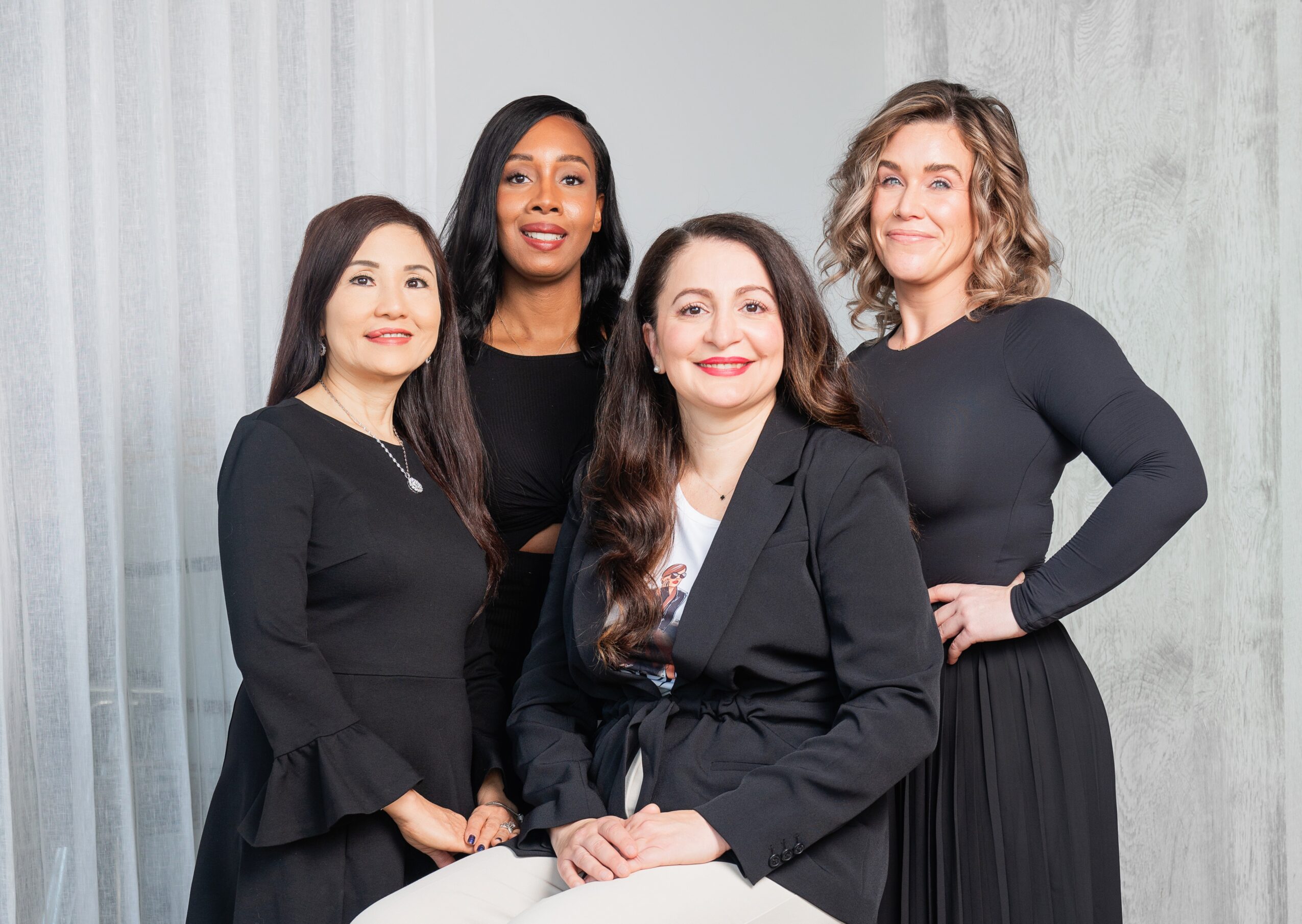 Four women standing together, smiling, in front of a light-colored curtain background.