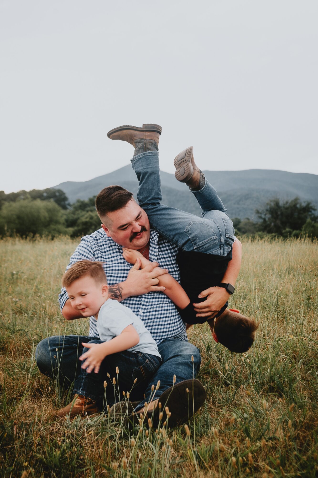 Three people playing in a grassy field with mountains in the background, one person upside down, two smiling children.