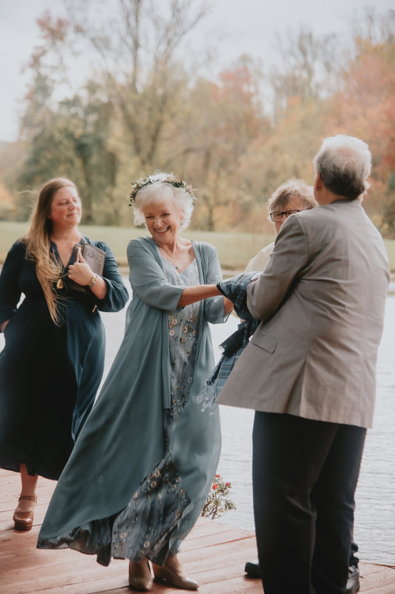 Group of people dancing outdoors near a body of water, trees in the background, woman in a long dress smiling.