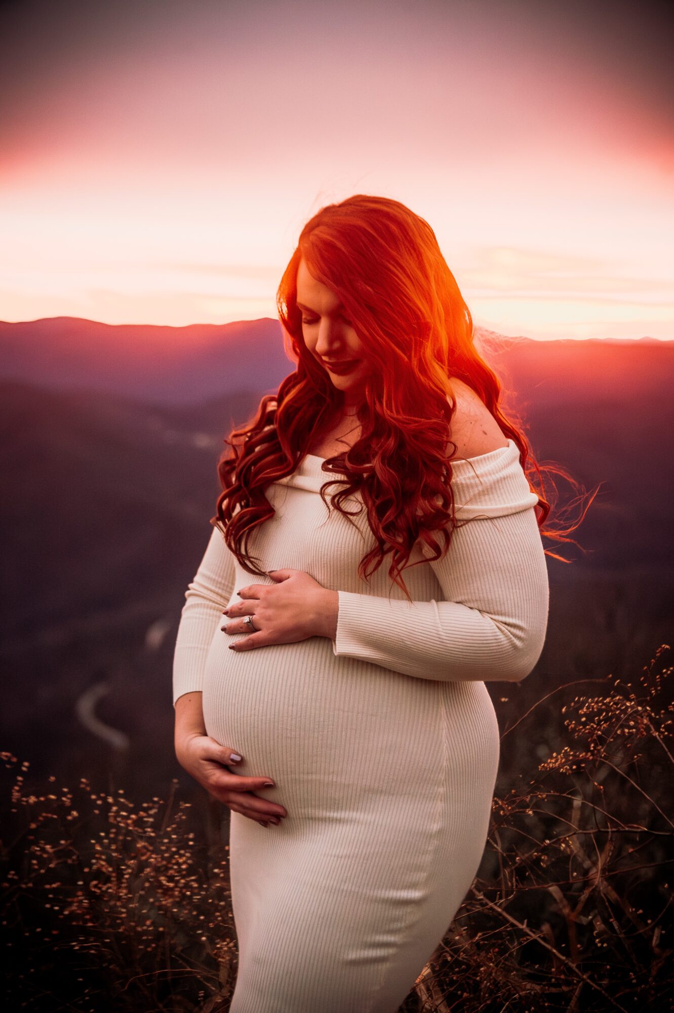Pregnant woman with long red hair standing outdoors at sunset, holding her belly with both hands, smiling gently.