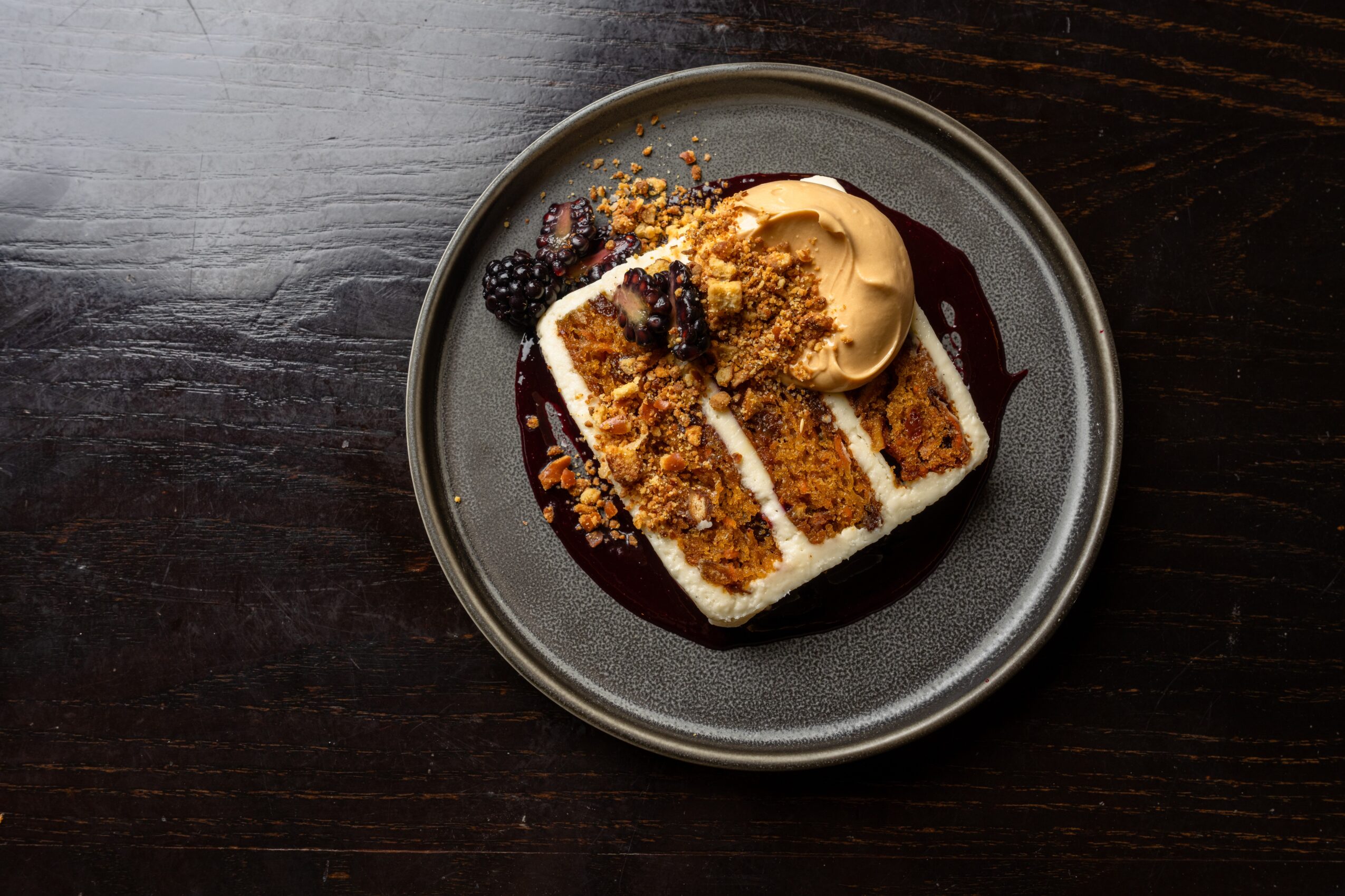 Slice of cake with cream, berries, and crumble on a dark plate, dark wooden background