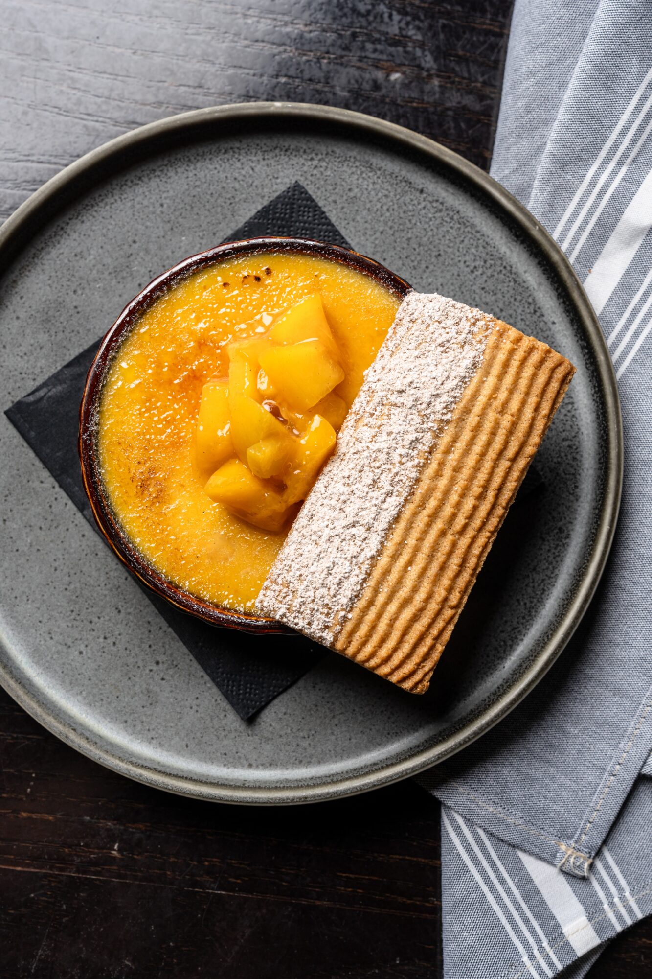 Dessert with mango topping and two layered cake slices on a dark plate, on a gray napkin, viewed from above.