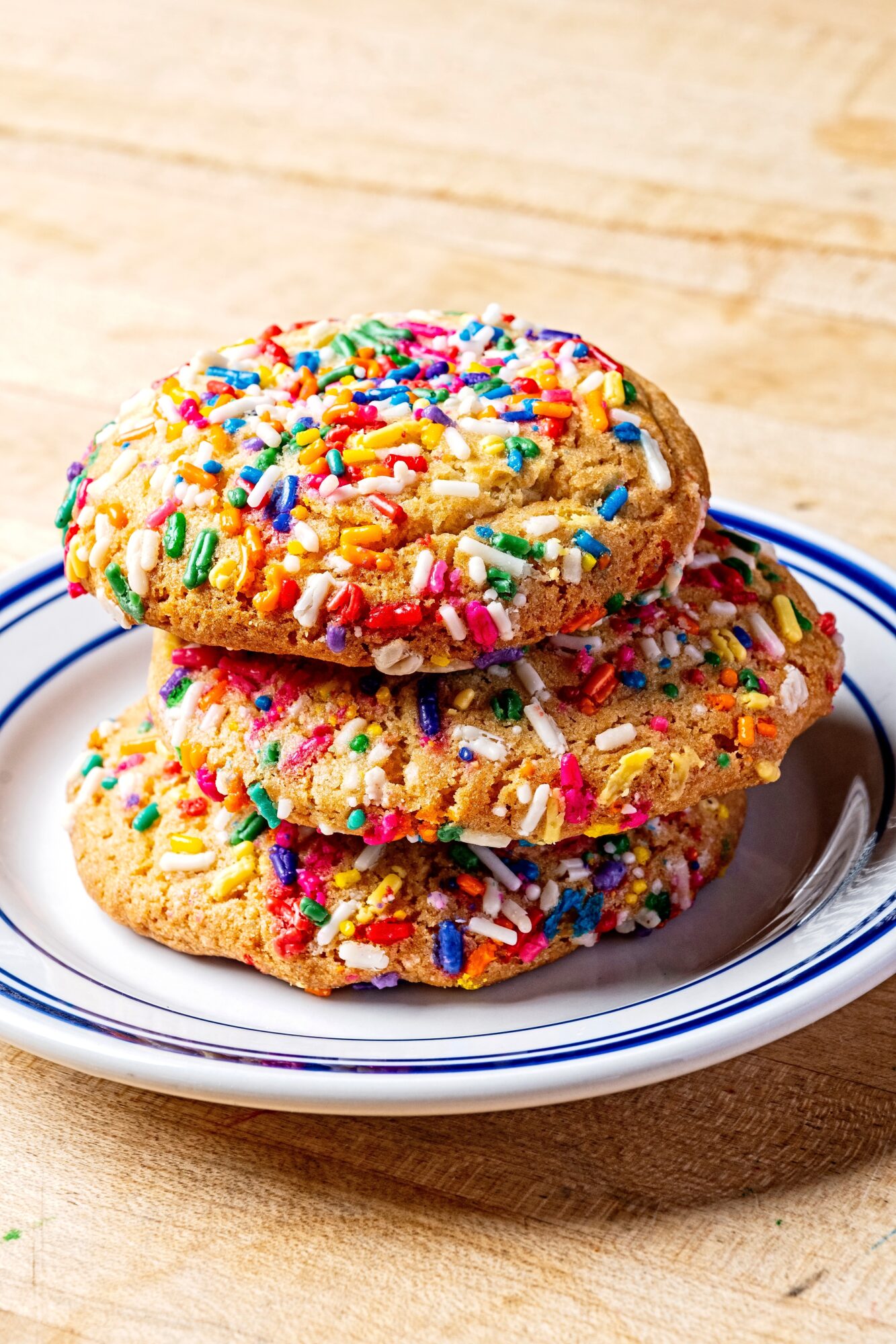 Three cookies with colorful sprinkles stacked on a white plate with blue rim, on a wooden surface.