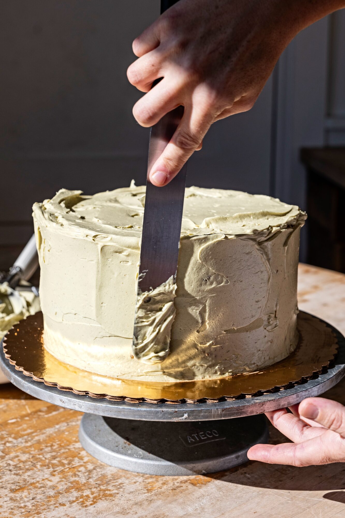 Person slicing a large cake with light-colored frosting on a cake stand, with a hand holding a knife.