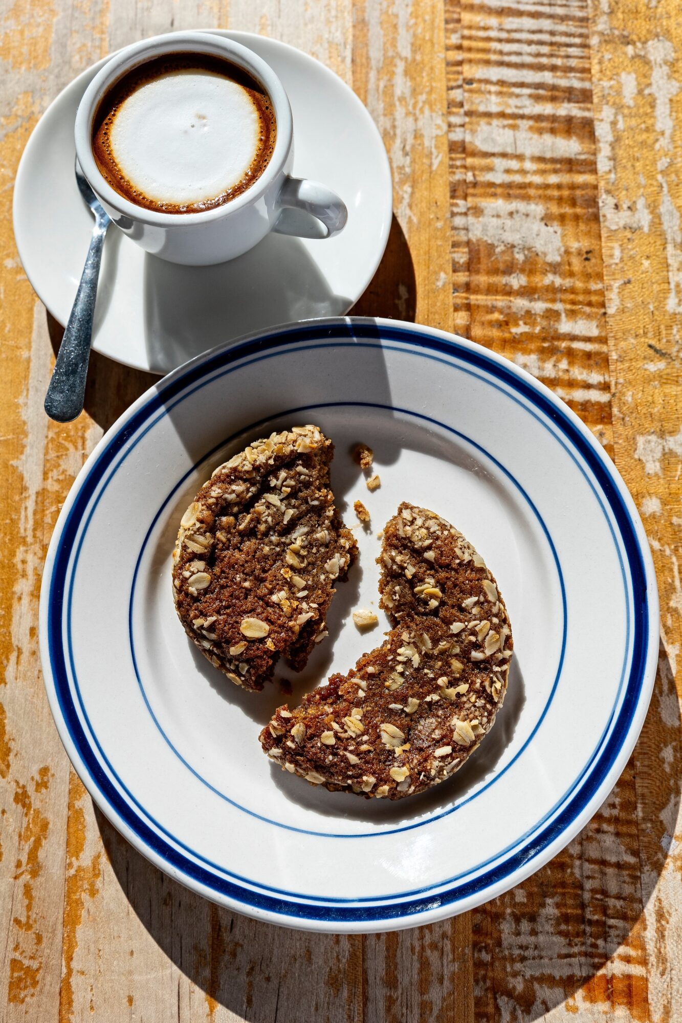 Cup of coffee with frothy top and a spoon, next to a sliced granola bar on a white plate with blue rim, on wooden table.