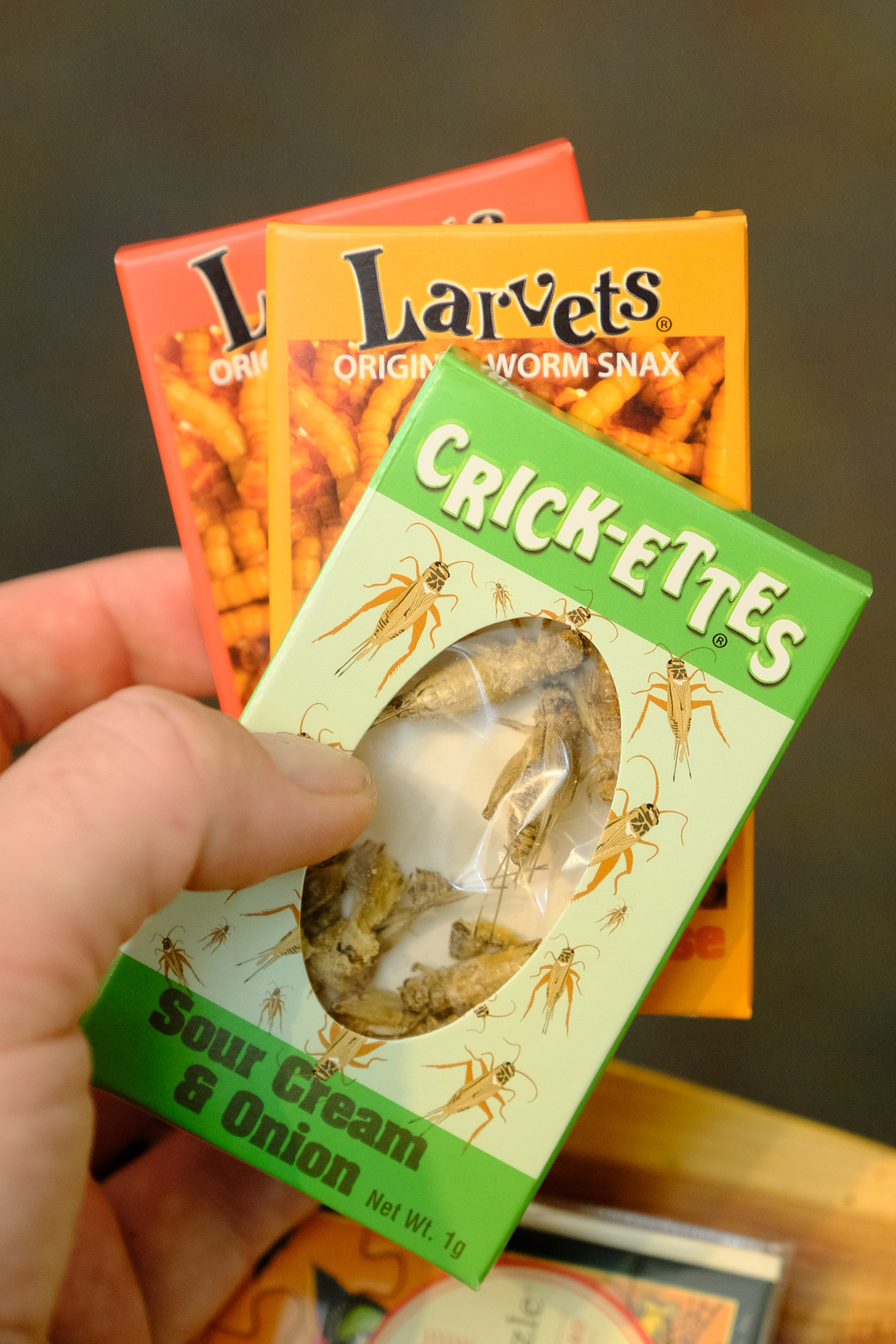 Hand holding a box of Crick-ettes sour cream and onion snack crackers, with other snack boxes in background.