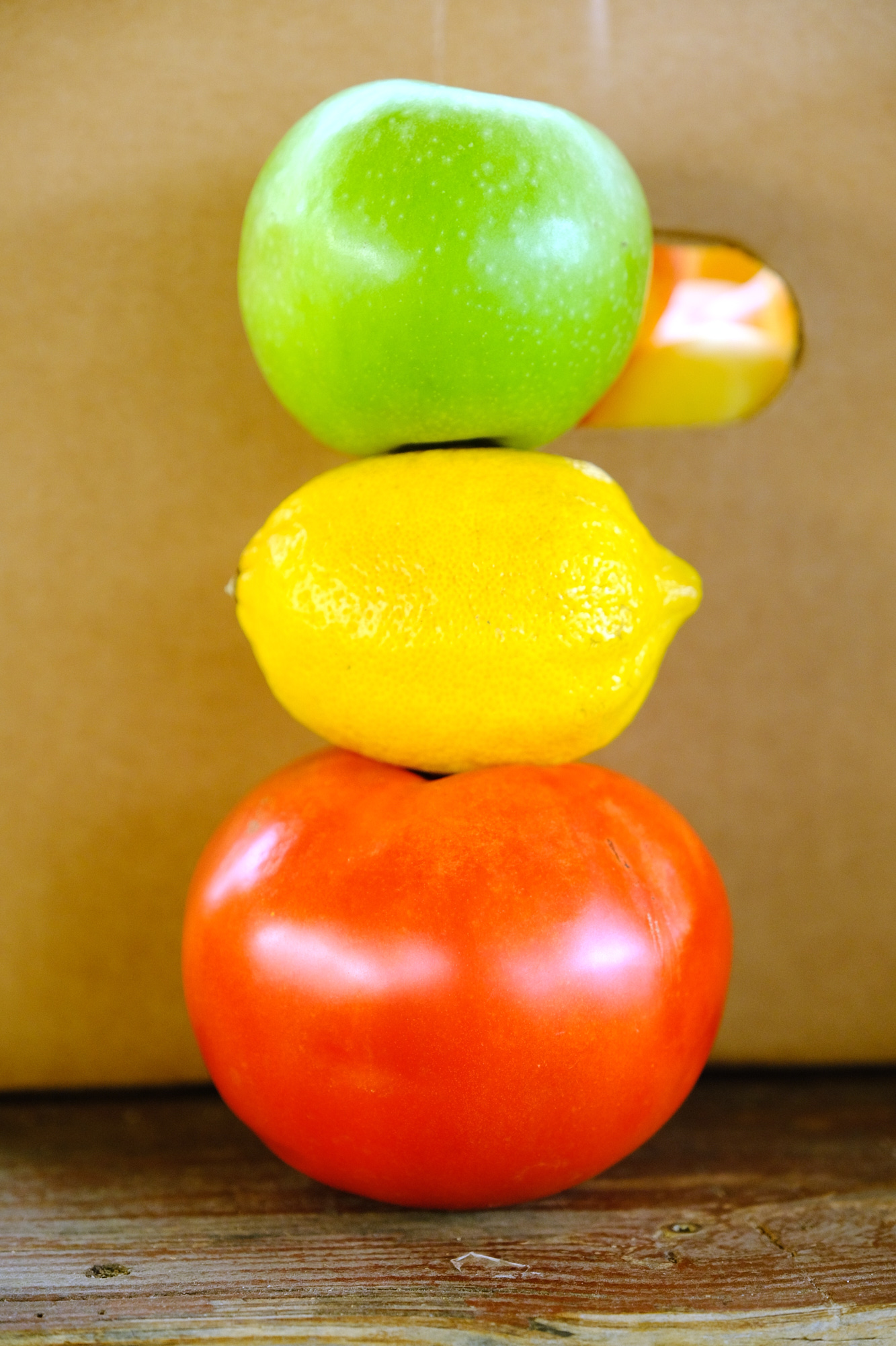 Stacked green apple, yellow lemon, and red tomato on a wooden surface.