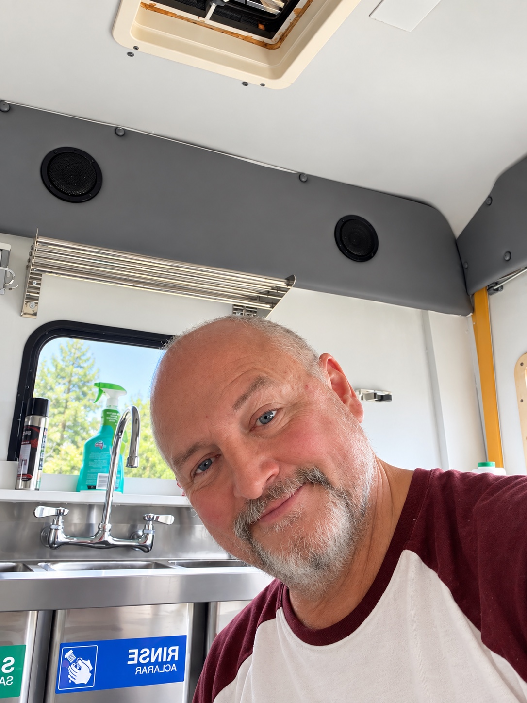 Man with a beard smiling inside a vehicle or trailer, with a window and a sink in the background.