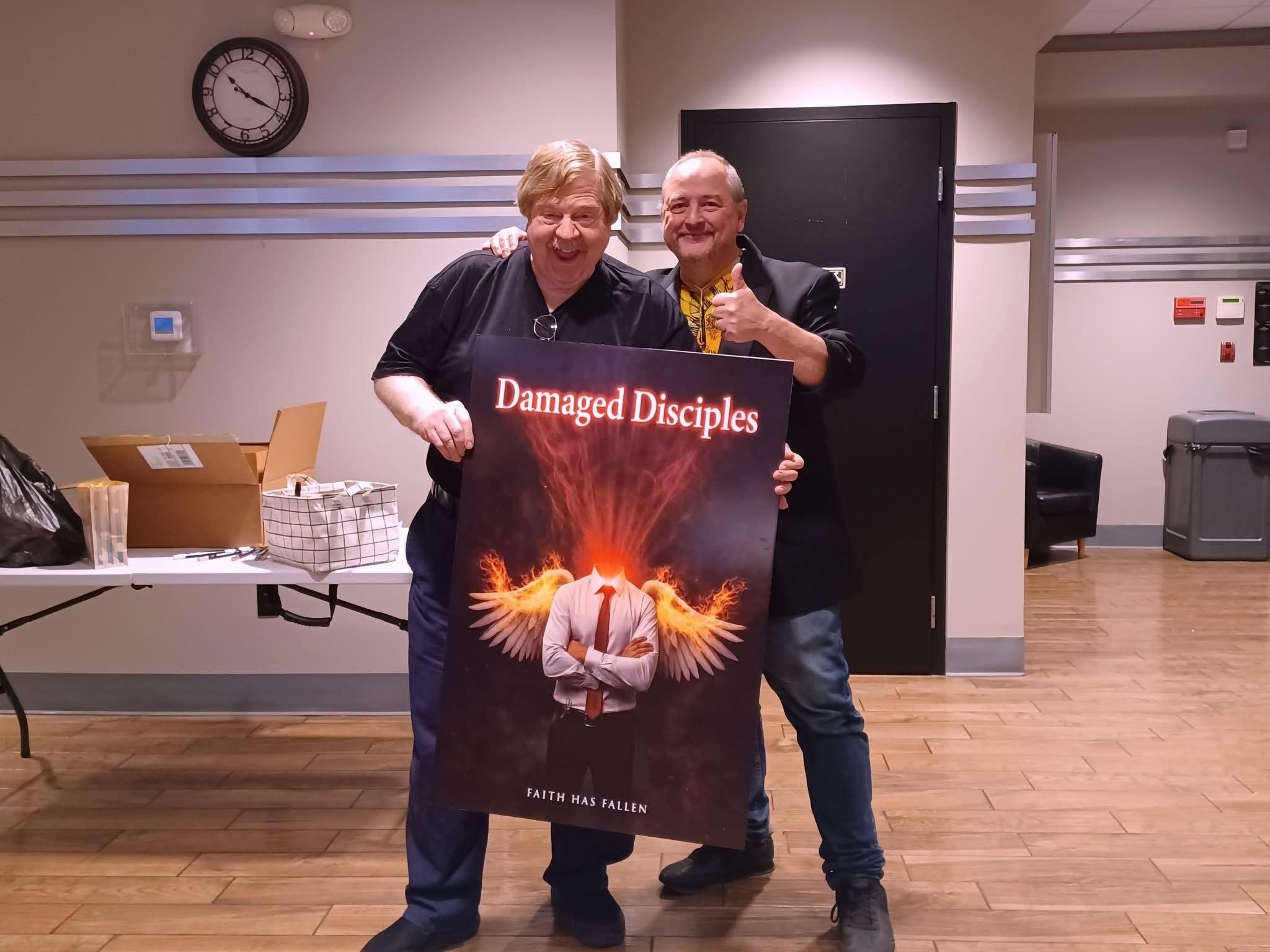 Two men holding a large poster with a person and fiery wings, smiling indoors with a clock on the wall.