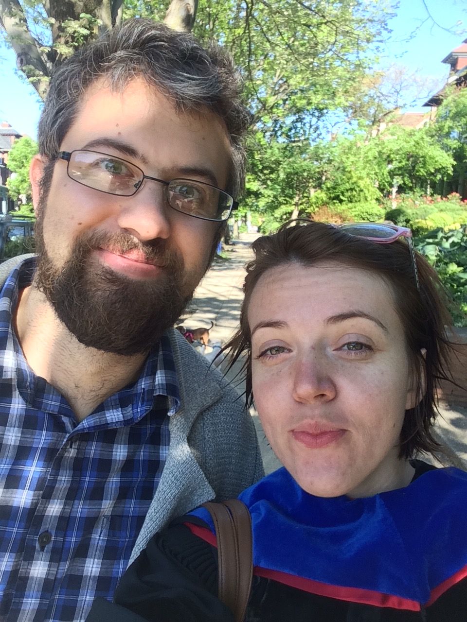 Two people outdoors, one with glasses and a beard, the other sticking out tongue, trees and sky in background.