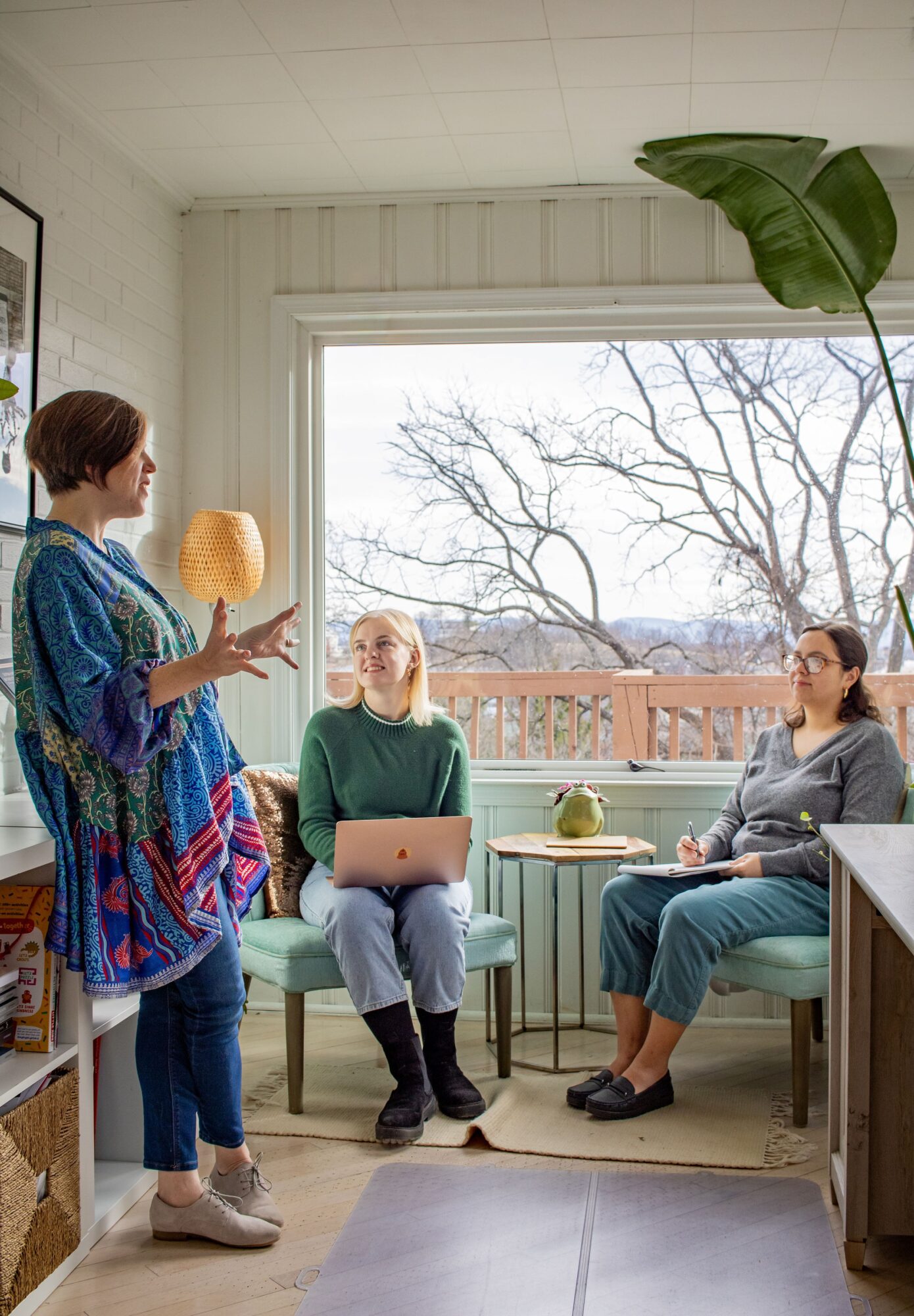 Three women in a room with a large window, one standing and two sitting, engaged in conversation.