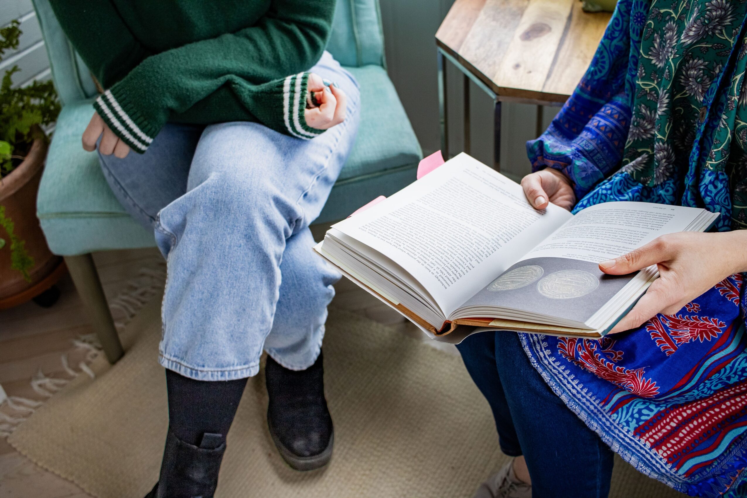 Two people sitting on a sofa, one holding an open book, the other with hands crossed, in a cozy room.