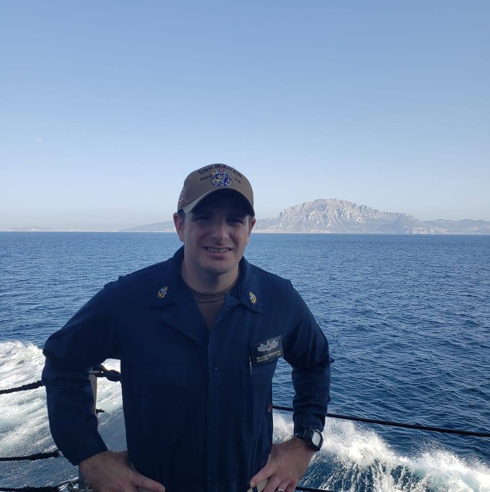 Person in uniform standing on a boat with ocean and distant land in background, smiling, daytime.