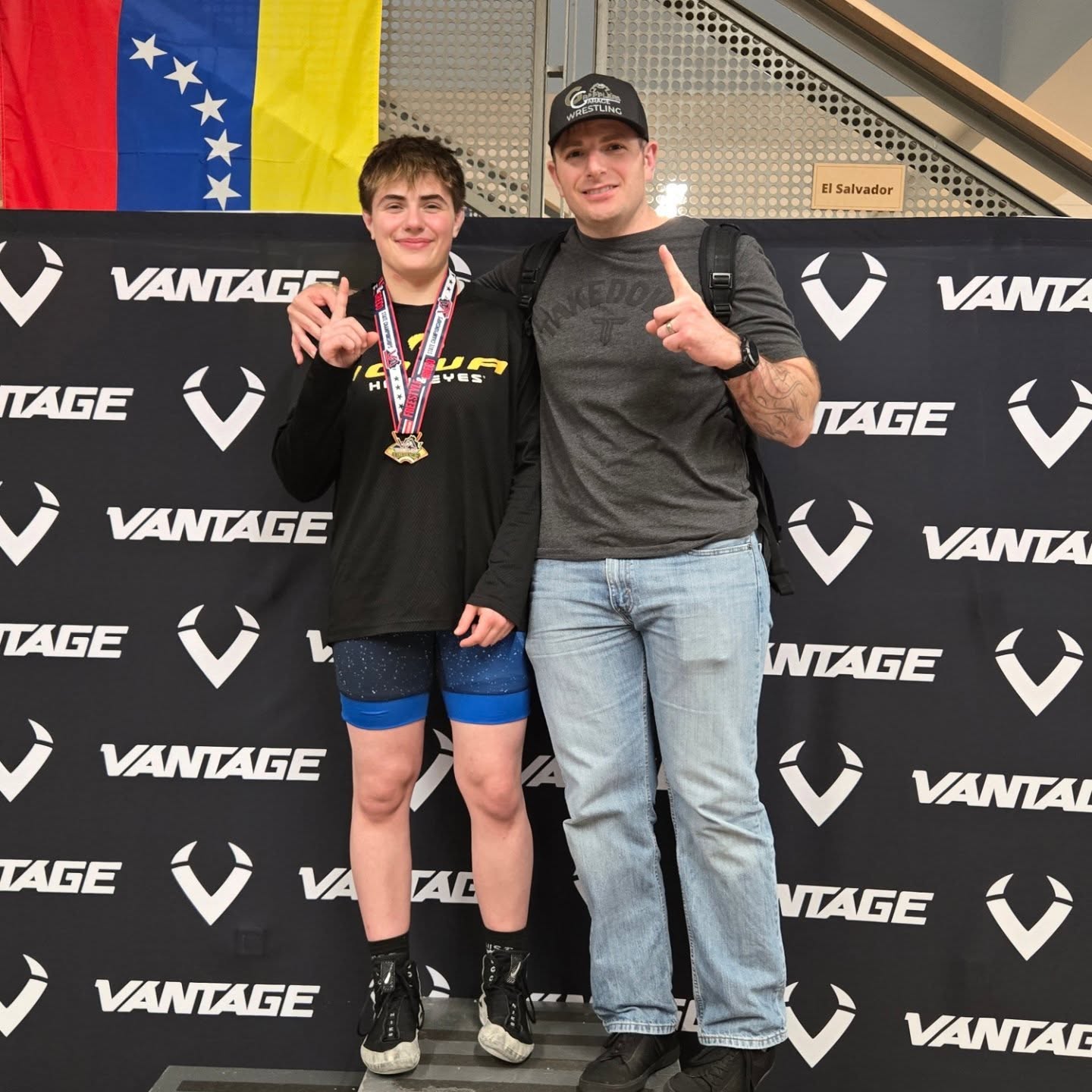 Two people standing in front of a black backdrop with VANTAGE logo, one with medals, Venezuelan flag in background.